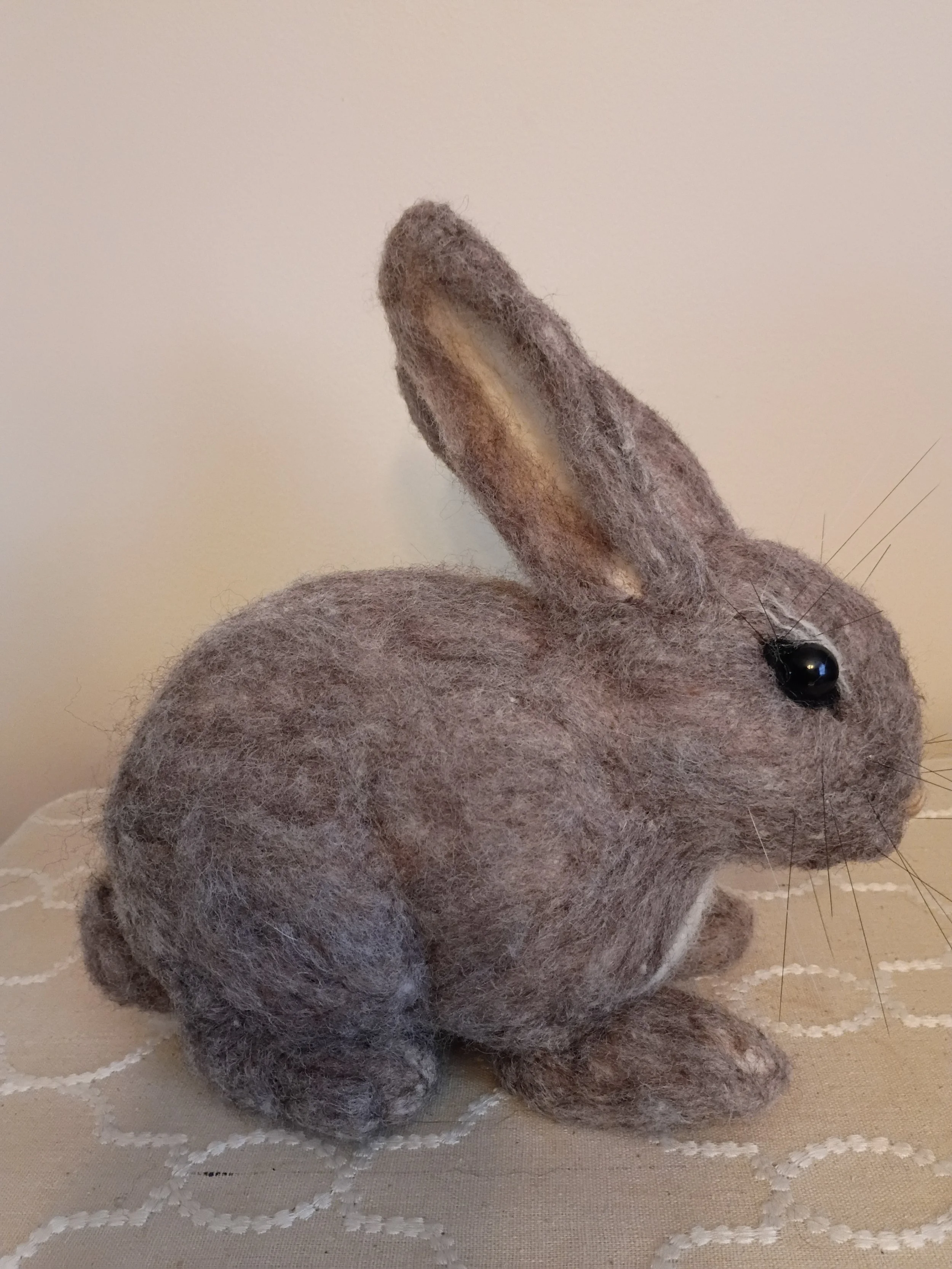 A felted gray stuffed mouse toy with black eyes, a long tail, sitting on a beige tablecloth with white stitched pattern, against a plain beige wall.