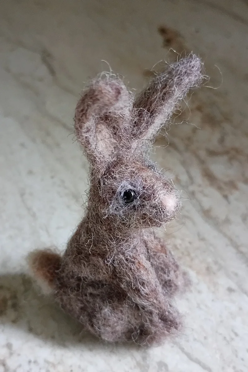 A small, felted animal resembling a bunny or rabbit with large ears, black eyes, and a woolly texture, sitting on a beige surface.