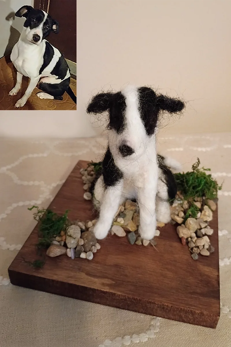 A felted wool sculpture of a black and white dog with a black patch over one eye, sitting on a wooden platform with small rocks and green moss around it, with a photograph of the real dog in the background.