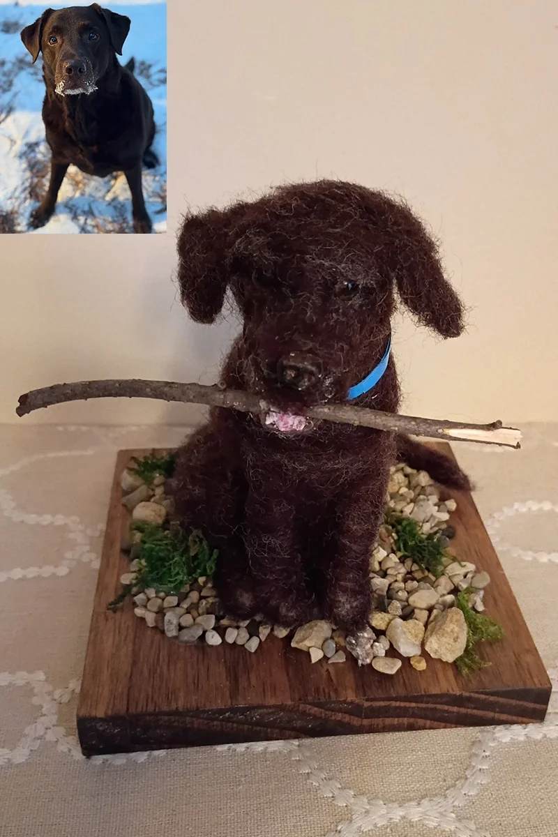 A small, handmade figurine of a brown dog with a rough, textured surface, sitting on a wooden base surrounded by small rocks and greenery, holding a stick in its mouth, with a blue collar around its neck.