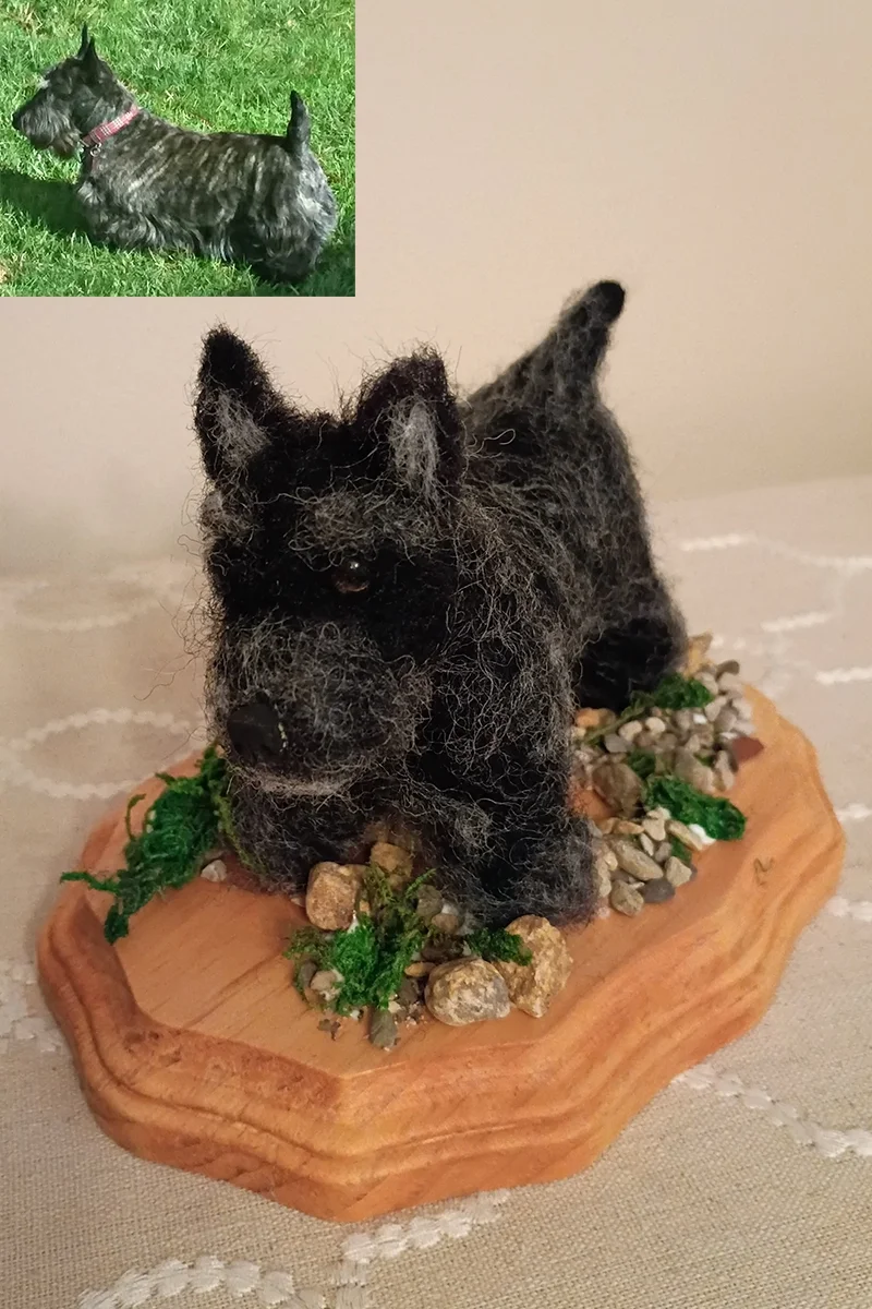 A miniature sculpture of a black dog with a fuzzy, textured coat, lying on a wooden base decorated with small rocks and green moss, with an inset photo of the real dog standing on grass in the top left corner.