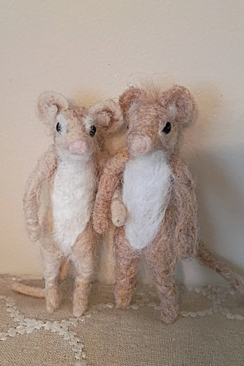 Two handcrafted felt mice figurines with pink noses, black eyes, and light brown and white fur, standing side by side against a plain background.