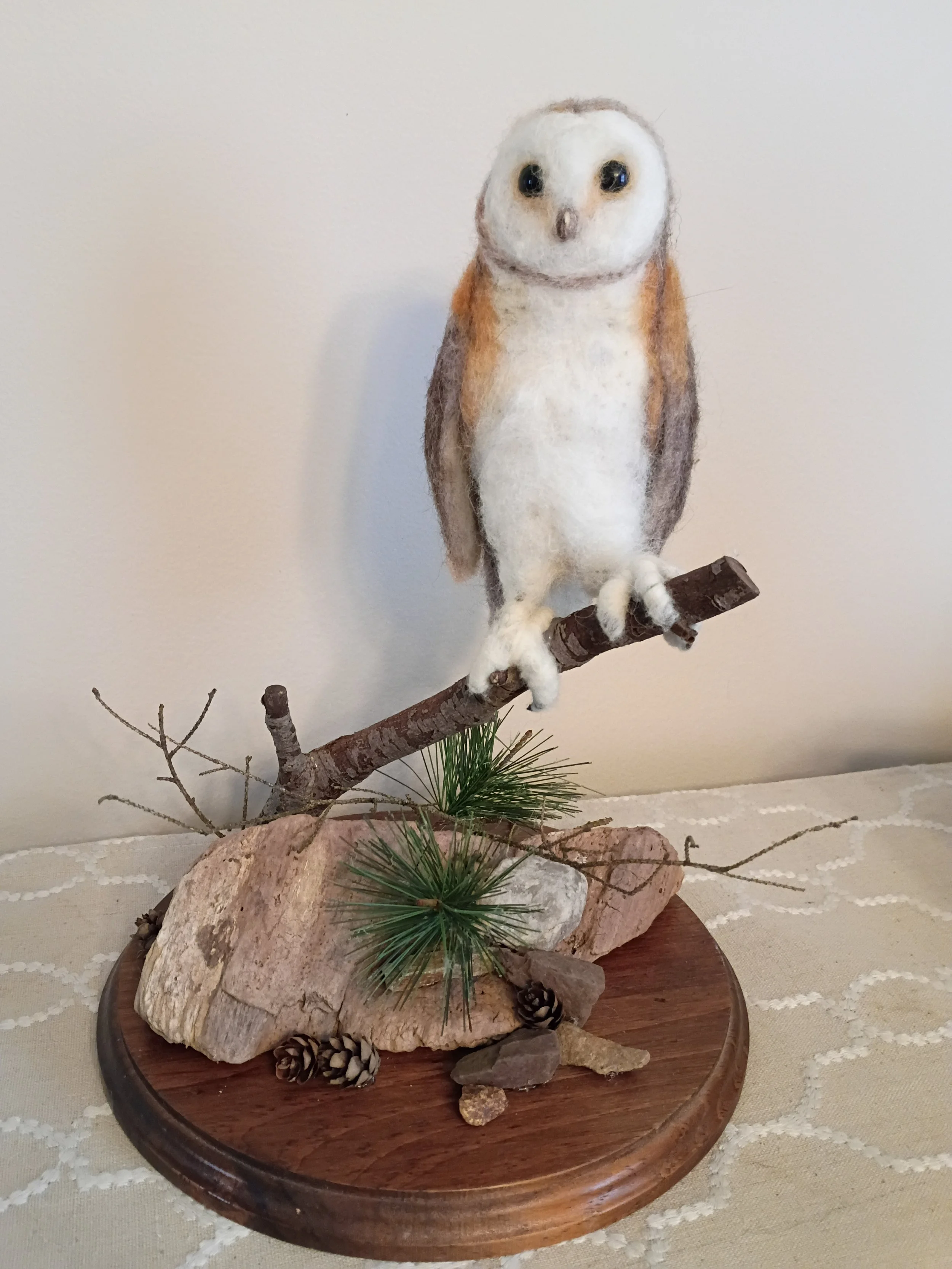 A felted owl figurine perched on a branch, surrounded by rocks, pinecones, and pine branches, on a round wooden base.
