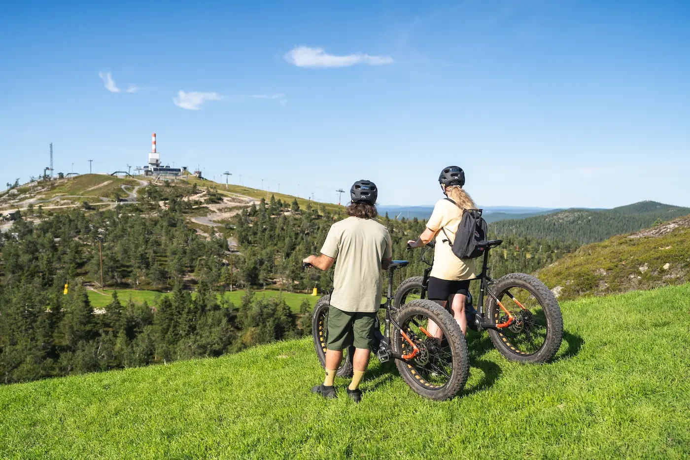Kaksi henkilöä katselee toisiaan ja polkee fatbike-pyörillä vuoristomaisemassa, taustalla on konemasto ja taivasta.