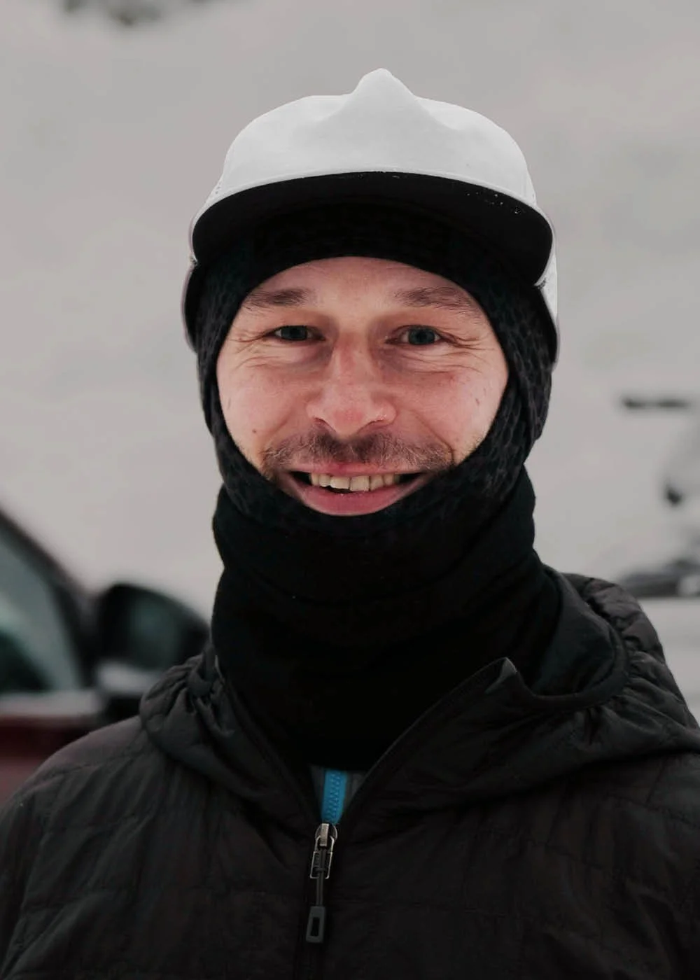 Henkilö hymyilee talvipuvussa, päässään lippalakki ja kauluri, taustalla autoja ja lumisadetta.