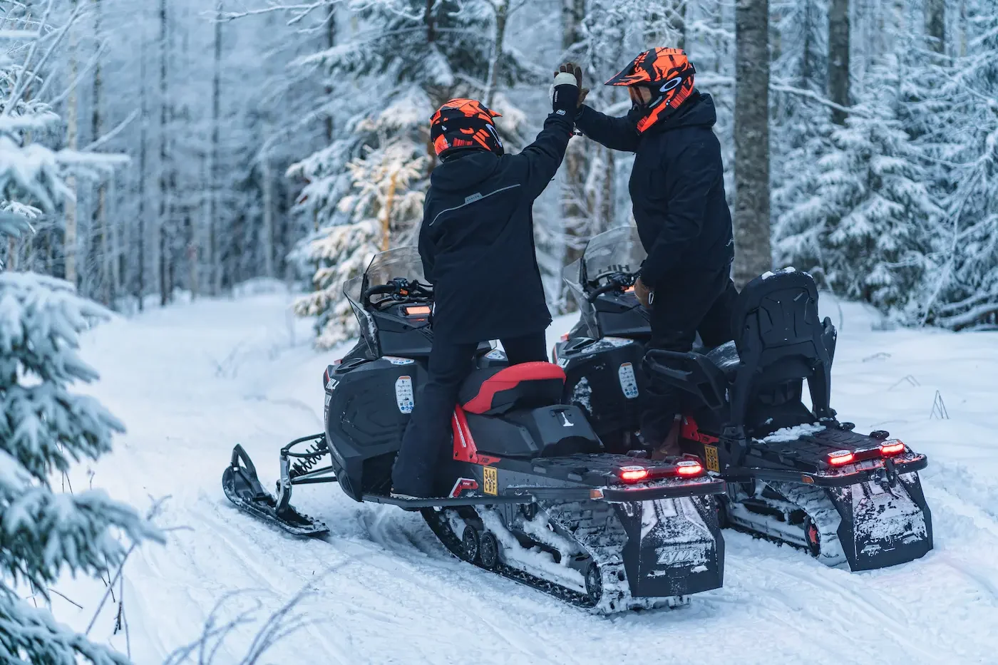 Kaksi ihmistä juttelemassa lumisella metsäreitillä moottorikelkoilla.