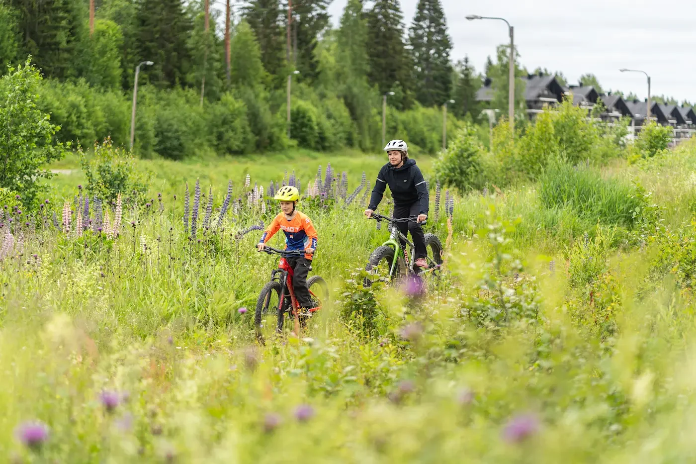 Aikuinen ja lapsi pyöräilevät luonnonkauniissa metsäisessä paikassa, ympärillään villiä kasvillisuutta ja kukkia.