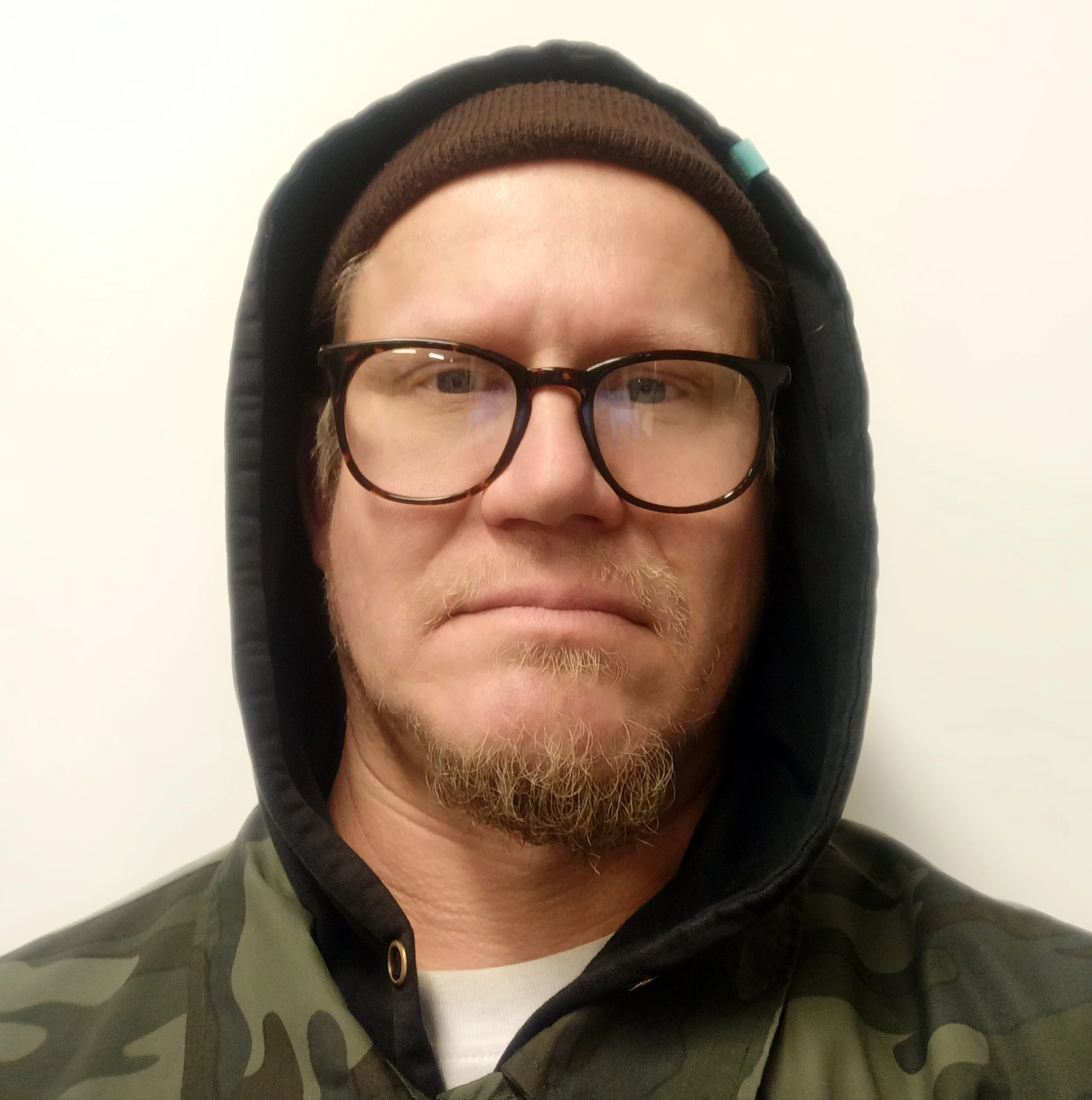 Lähikuva miehestä, jolla on ruskeat silmälasit, pipo ja takki, taustalla valkoinen seinä.