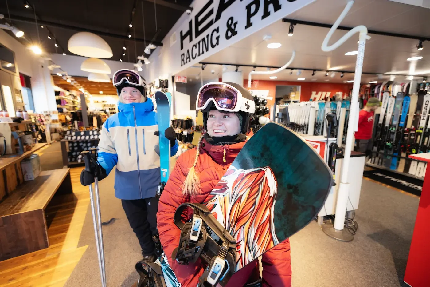 Kaksi nuorta naista shoppailemassa urheiluvälineitä, toinen kantaa lumilautaa ja toinen pipoja ja ski-laseja, sisätiloissa urheiluvälinemyyntiliikkeessä.
