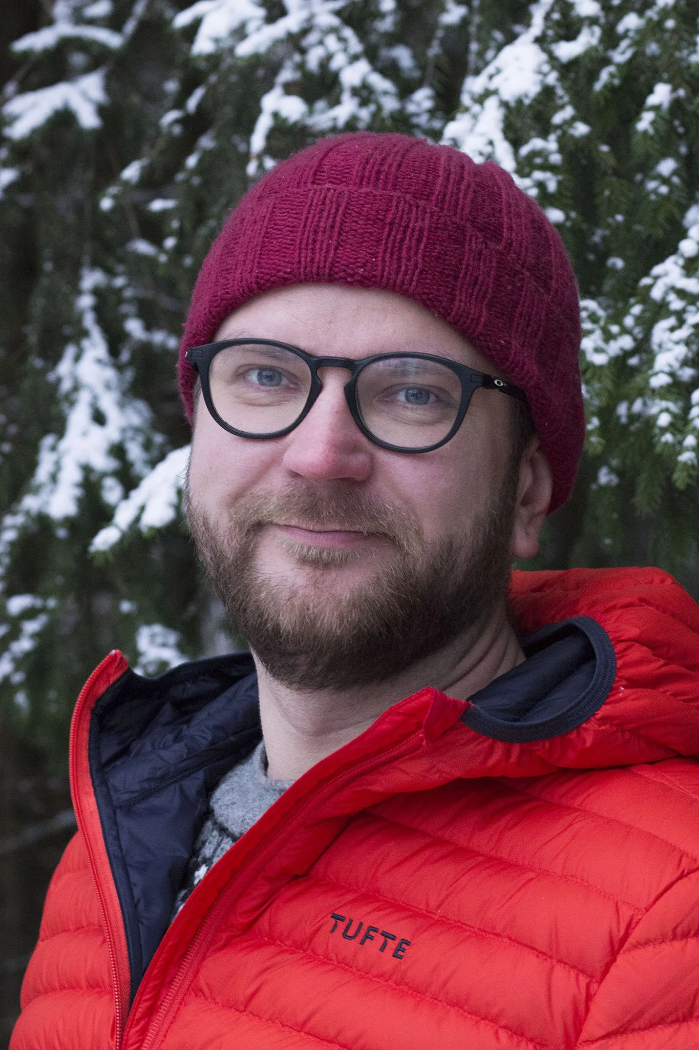 Mies hymyilee ulkona talvella, pukeutuneena punaiseen takkiin ja mustiin silmälaseihin, taustalla lumisia puita.