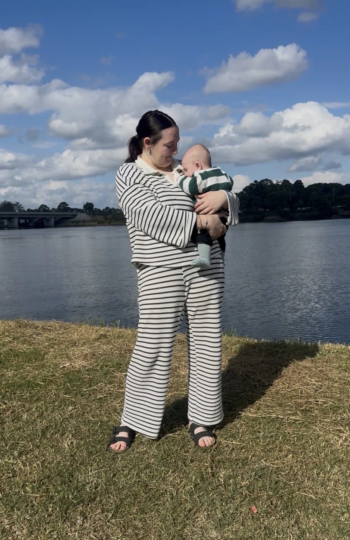 A woman holding a baby near a body of water on a sunny day with blue sky and clouds in the background.