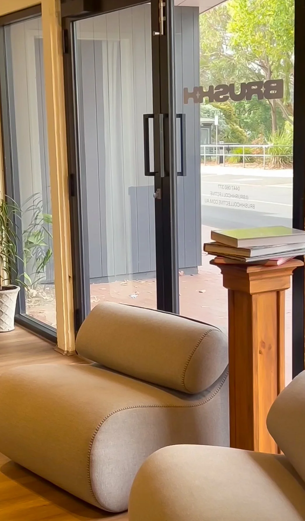 Inside a waiting area of a business, with a beige curved sofa, a wooden table with stacked books, glass doors with a BrushedH logo, and a view of greenery outside.