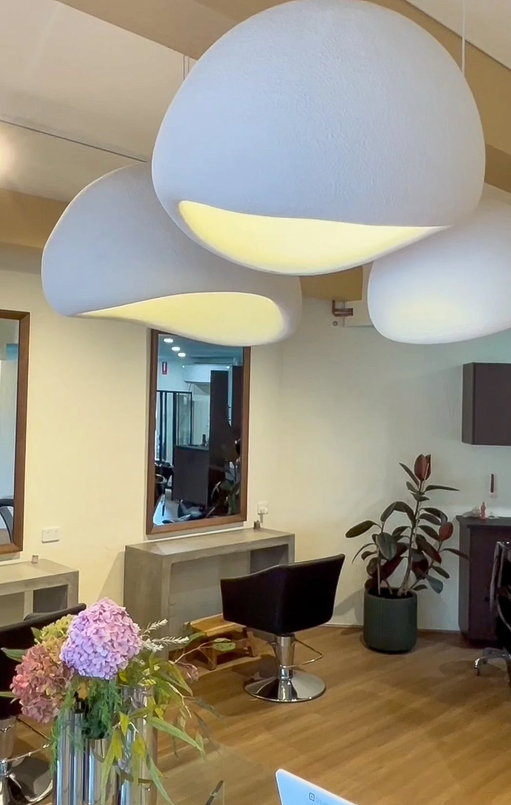 Modern interior space with large, abstract white ceiling lights, a wooden table with pink hydrangeas, a black salon chair, a mirror, a potted plant, and minimal decor.