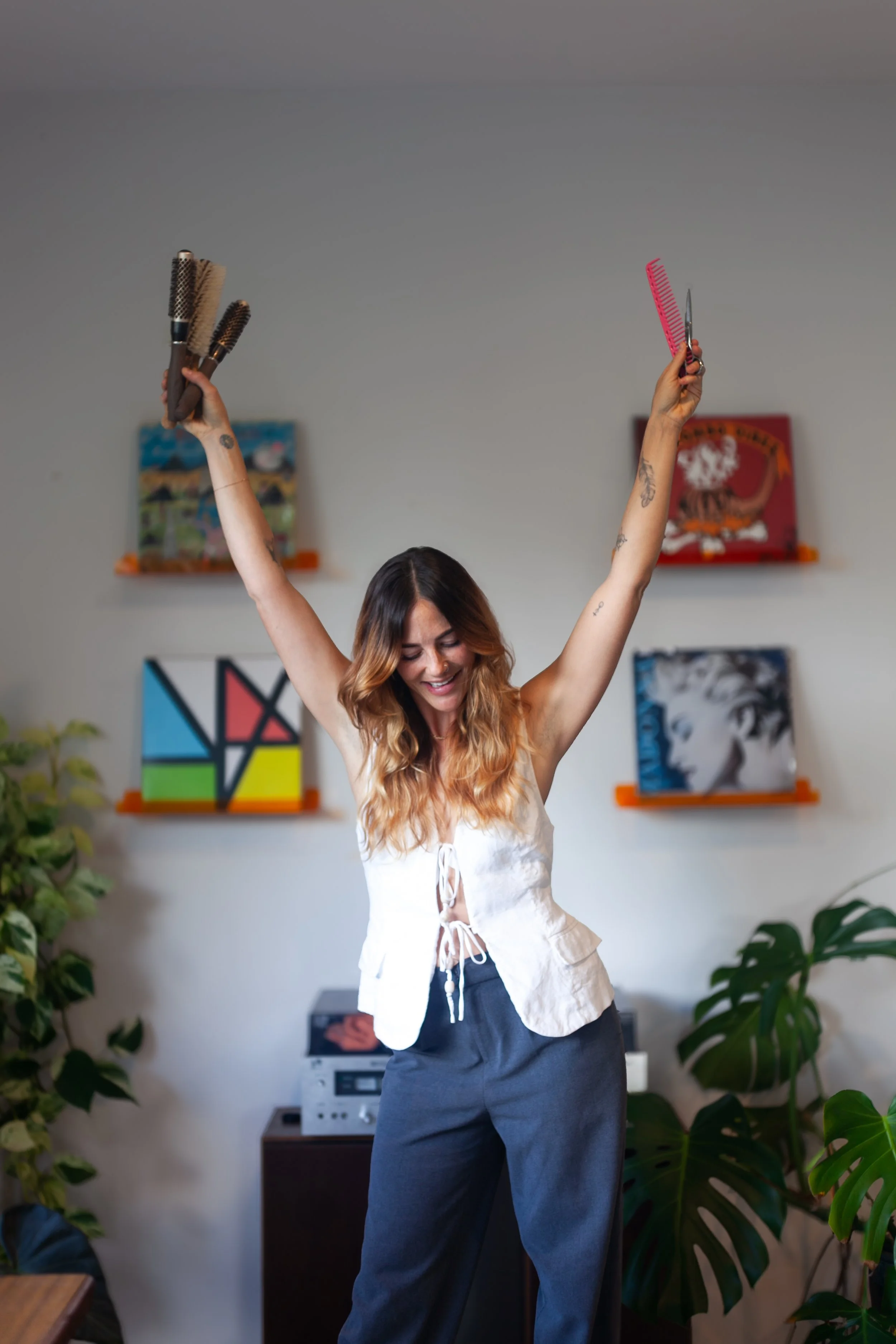A woman with long, wavy hair and tattoos on her arms, smiling with arms raised, holding hairbrushes in her left hand and a comb in her right hand, in a room with artwork on the wall and large green plants.