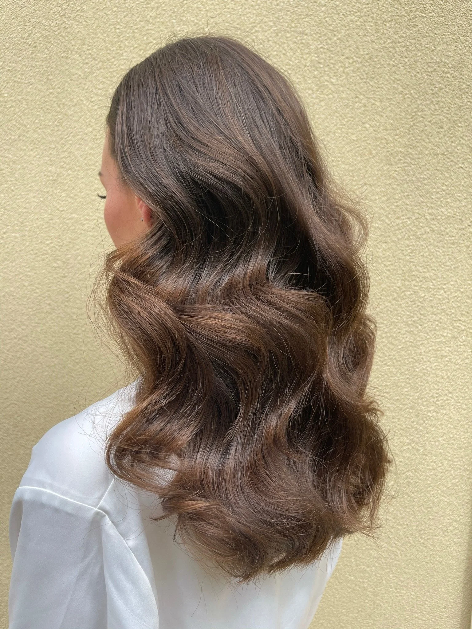 A woman with long, wavy, brown hair seen from the side, wearing a white shirt against a textured beige wall.