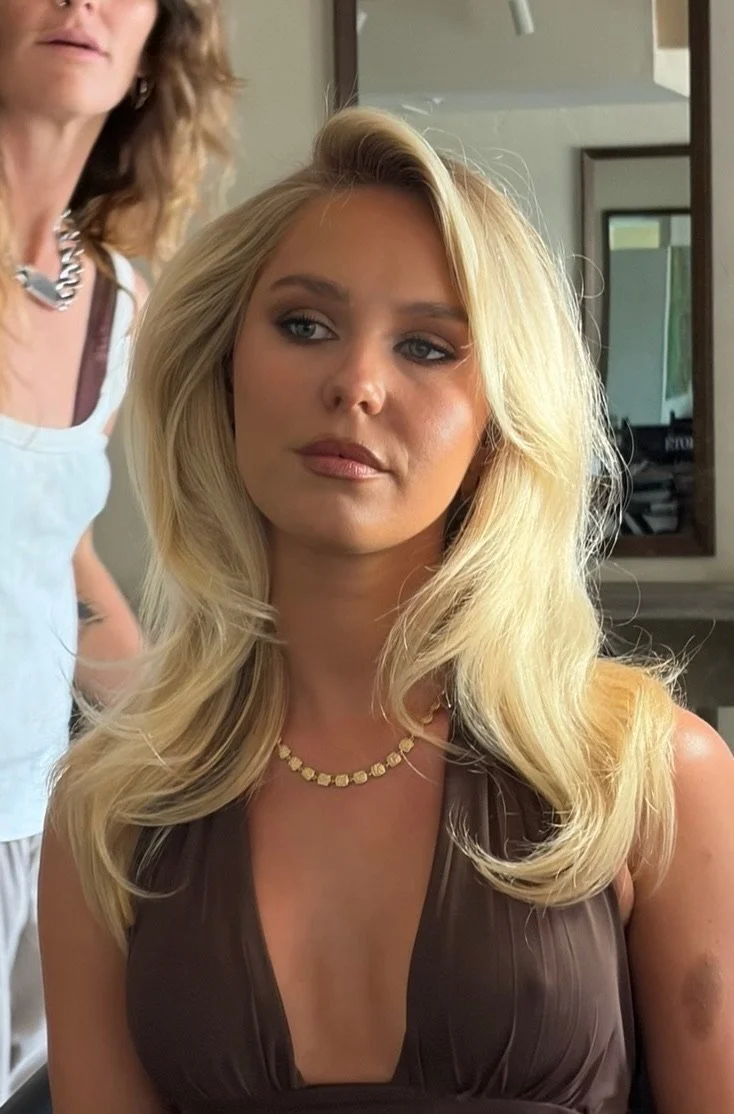 A woman with long blond hair styled in loose waves sits in a salon chair, wearing a brown dress and a gold necklace, looking off to the side. A stylist with curly hair and a white apron is partially visible behind her.