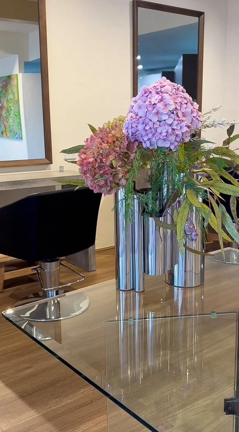 A bouquet of pink hydrangeas and other greenery in metallic vases placed on a glass table in a modern interior space.