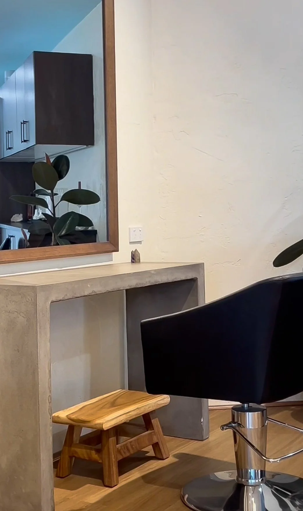 A modern interior space with a large mirror, black salon chair, small wooden stool, and a concrete vanity, reflected in the mirror is a kitchenette with black cabinets, a potted plant, and a wall outlet.