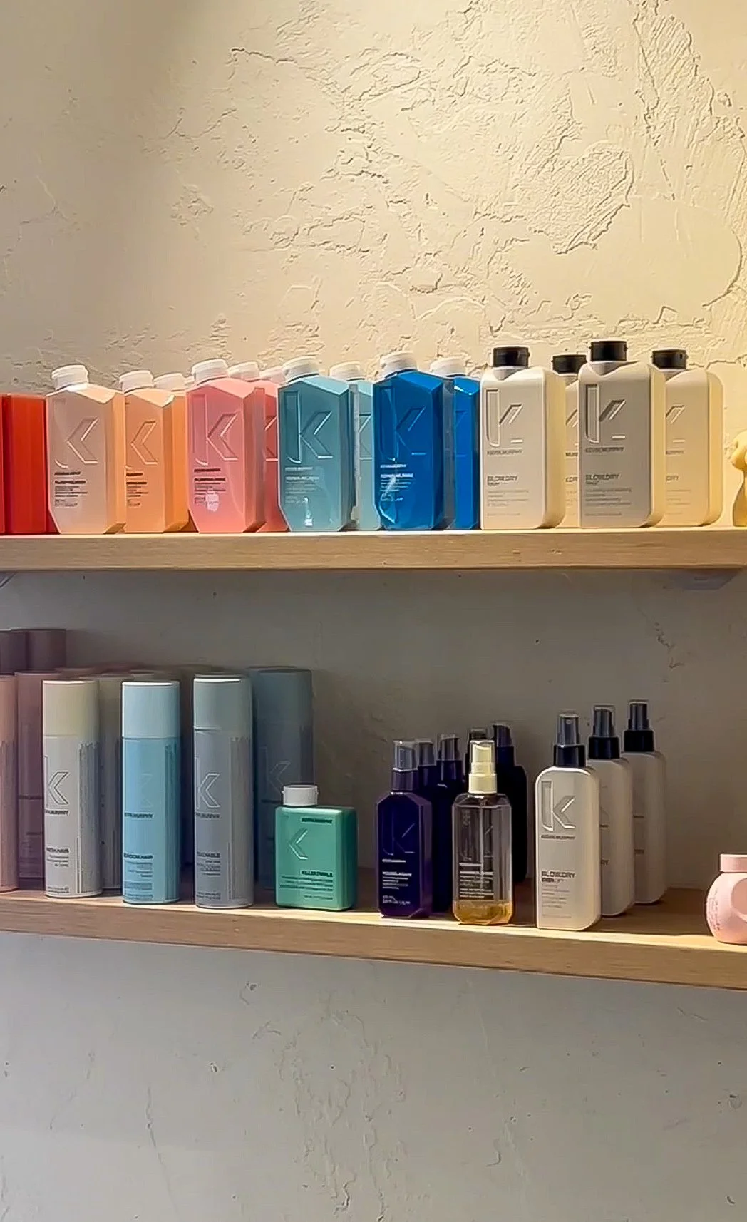 Shelf with colorful bottles of hair care products arranged in rows on two wooden shelves against a textured beige wall.