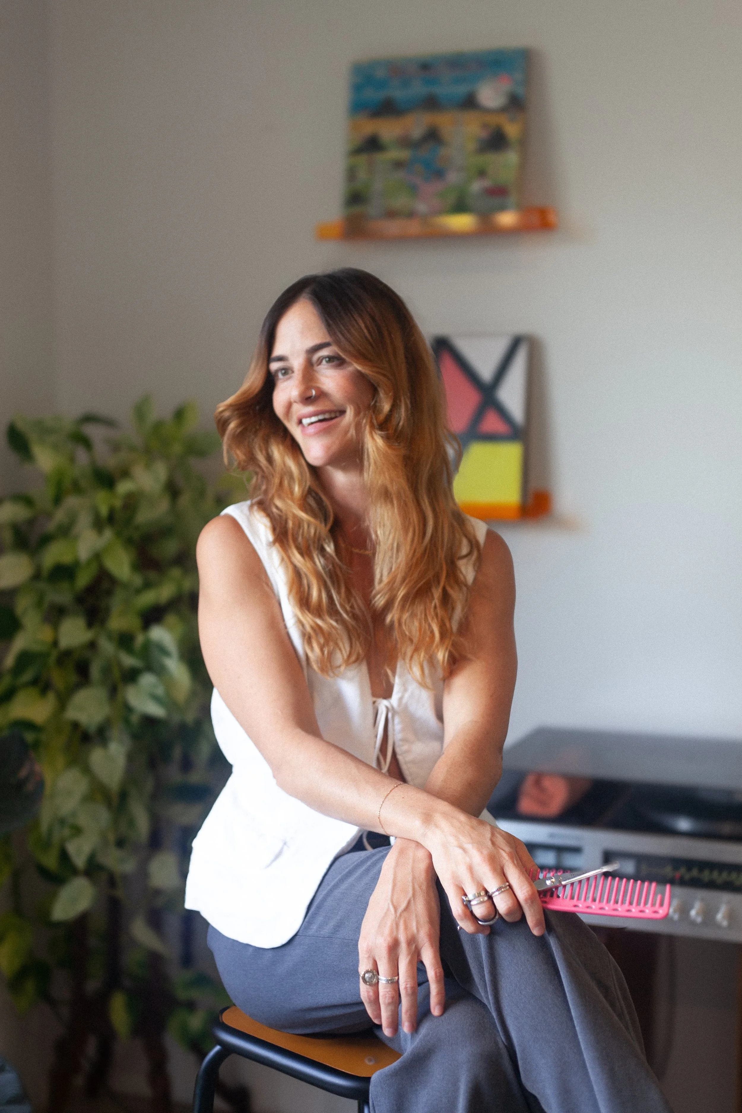A woman with long, wavy brown hair, wearing a white sleeveless top, sitting on a chair, smiling and looking to her right. She has rings on her fingers and a nose piercing. In the background, there are plants, and two colorful abstract paintings on the wall.