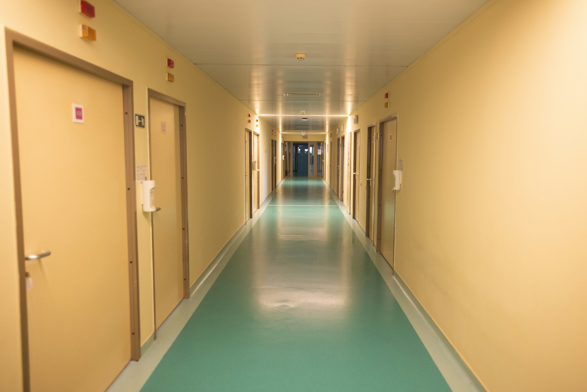 Long hospital corridor with yellow walls, green and white flooring, multiple closed doors on both sides, and ceiling lights.