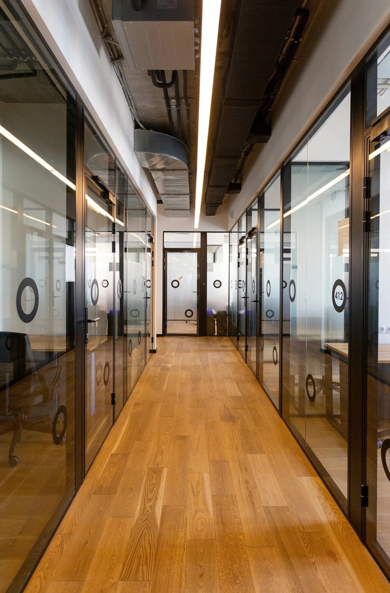 A corridor with glass walls and doors on both sides, displaying numbers and circular designs, with a wooden floor and a ceiling with exposed ductwork and pipes.