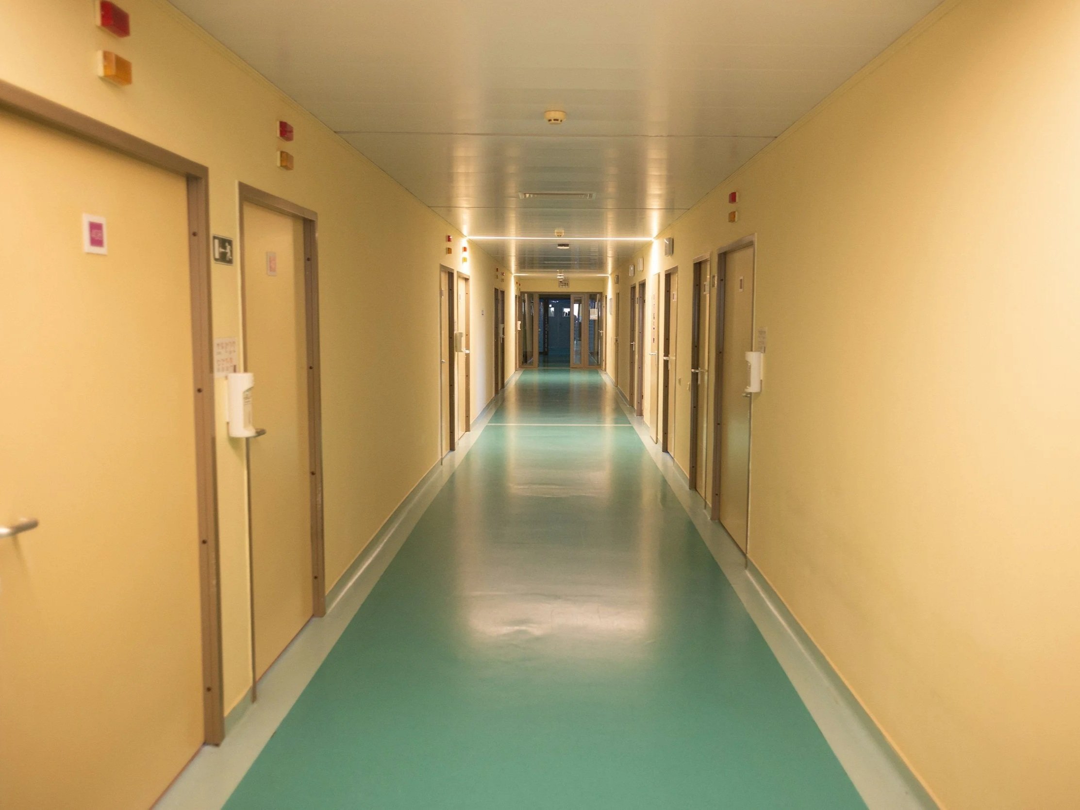A hospital hallway with beige walls and a patterned green and white floor, lined with closed doors and emergency equipment, leading to glass double doors in the distance.