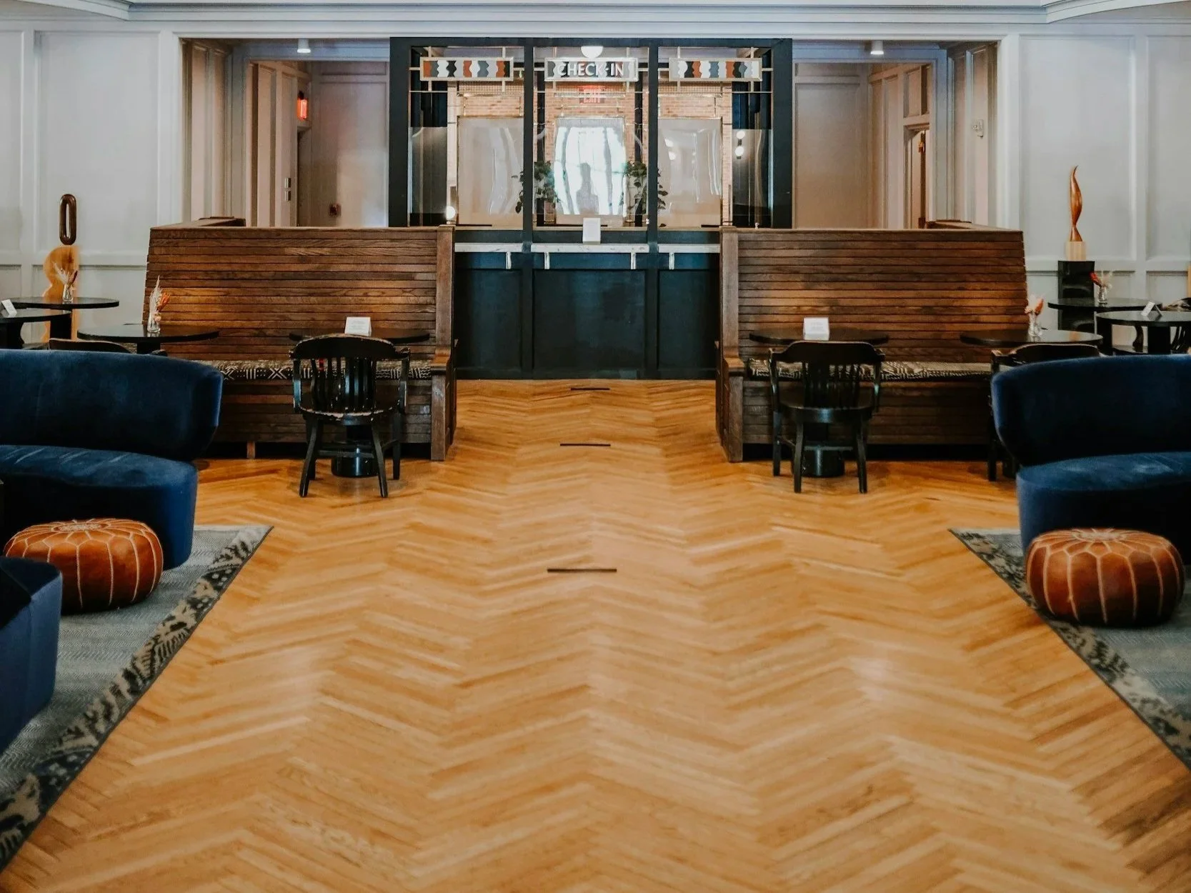 Interior of a modern hotel lobby with wooden flooring, seating area with blue sofas and orange ottomans, walls with decorative artwork, and a reception desk in the background.