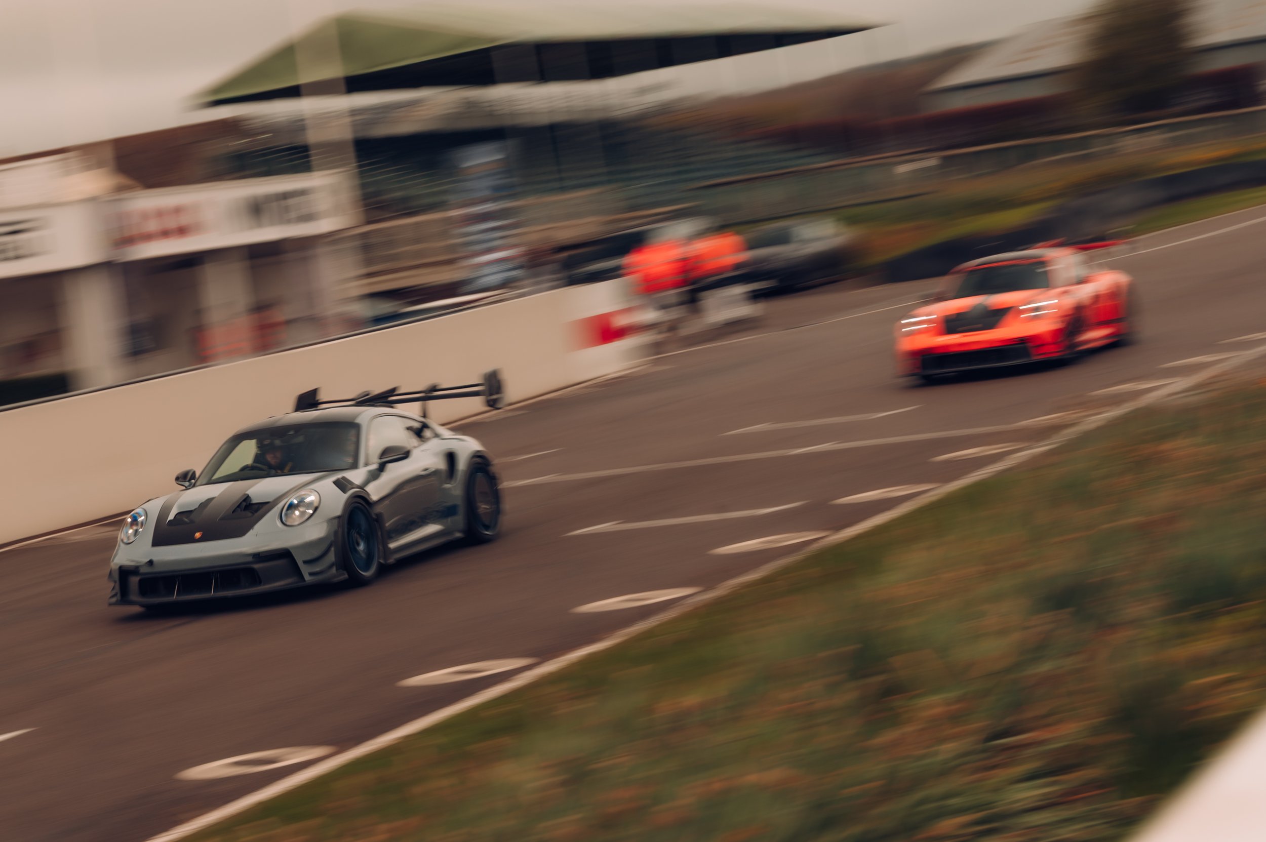 A silver sports car with black stripes and a rear spoiler racing on a track, with a red sports car in the background and a building with glass windows.