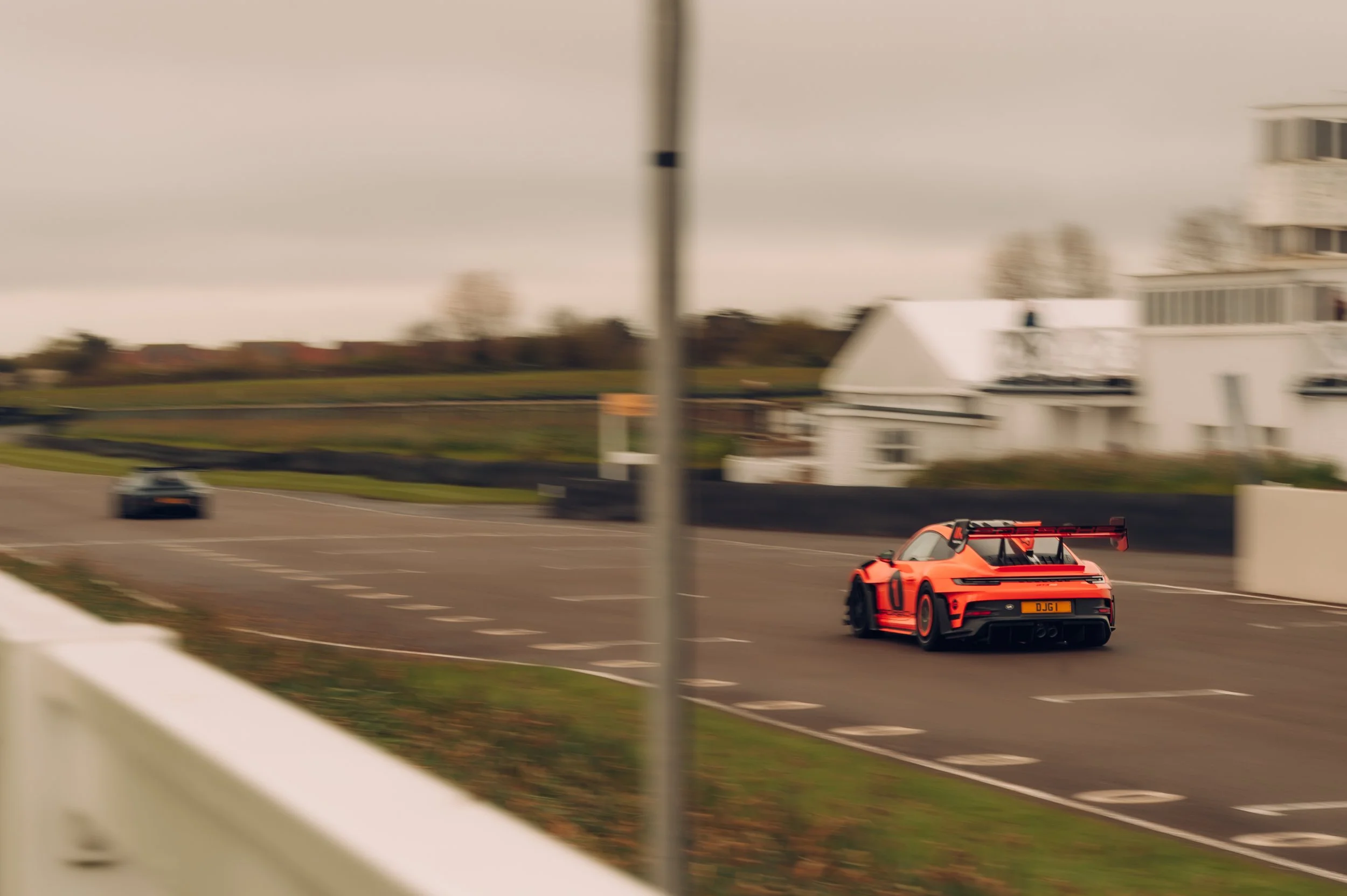 A bright orange race car driving on a track with a building and trees in the background during overcast weather.