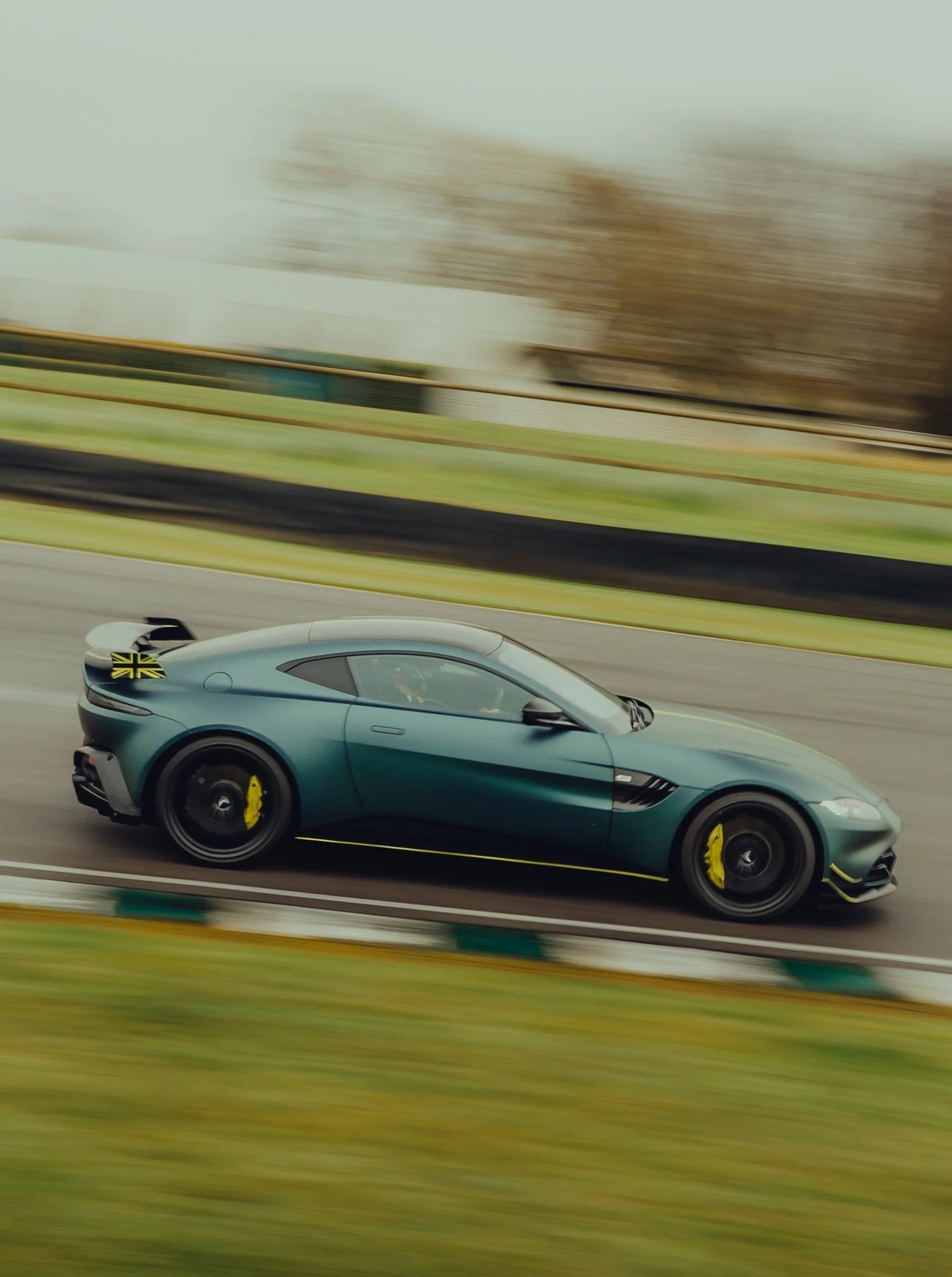 A silver sports car with black tires and yellow brake calipers driving on a highway with green grass and trees in the background, captured in motion.