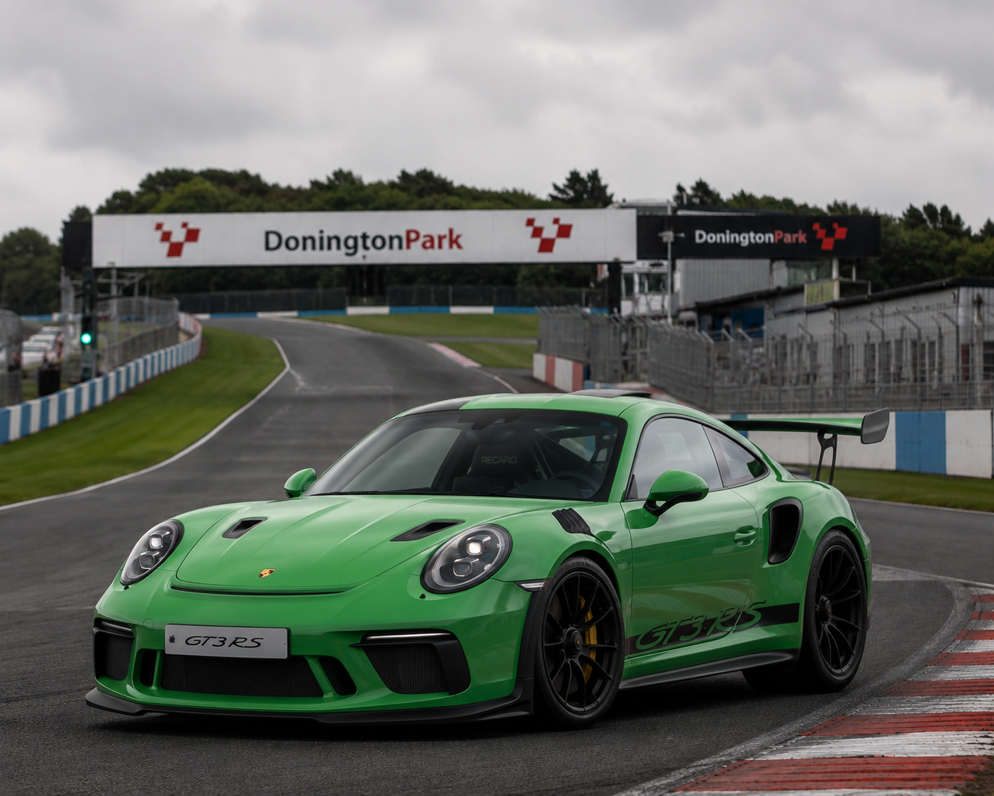 A bright green Porsche 911 GT3 RS race car on a racing circuit at Donington Park. The car features a large rear wing and black accents, with the model name visible on the front license plate.
