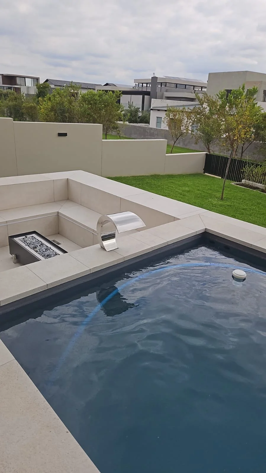 Modern backyard with a swimming pool, built-in seating area, and a fire feature, surrounded by a lawn, trees, and contemporary houses under cloudy sky.