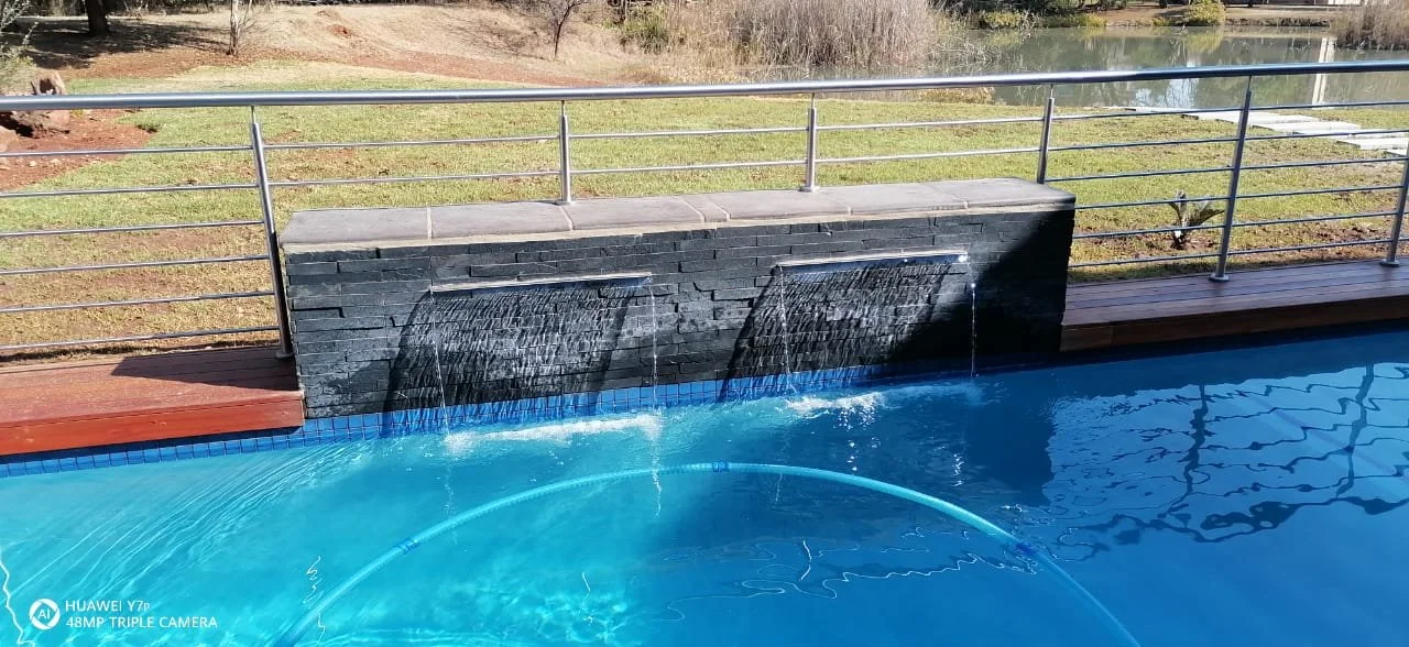 A swimming pool with water fountains cascading over a dark stone wall.