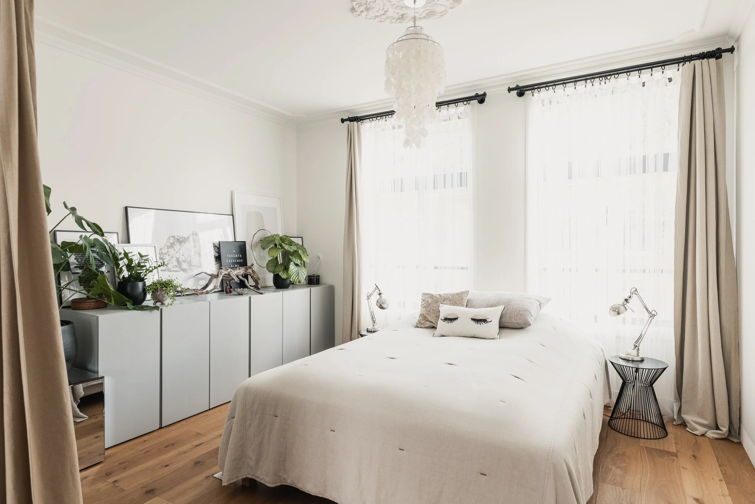A bright bedroom with white walls, large windows with sheer curtains, a bed with a white duvet and decorative pillows, two small black bedside tables with silver lamps, a gray sideboard decorated with books and plants, a ceiling chandelier, and wood 