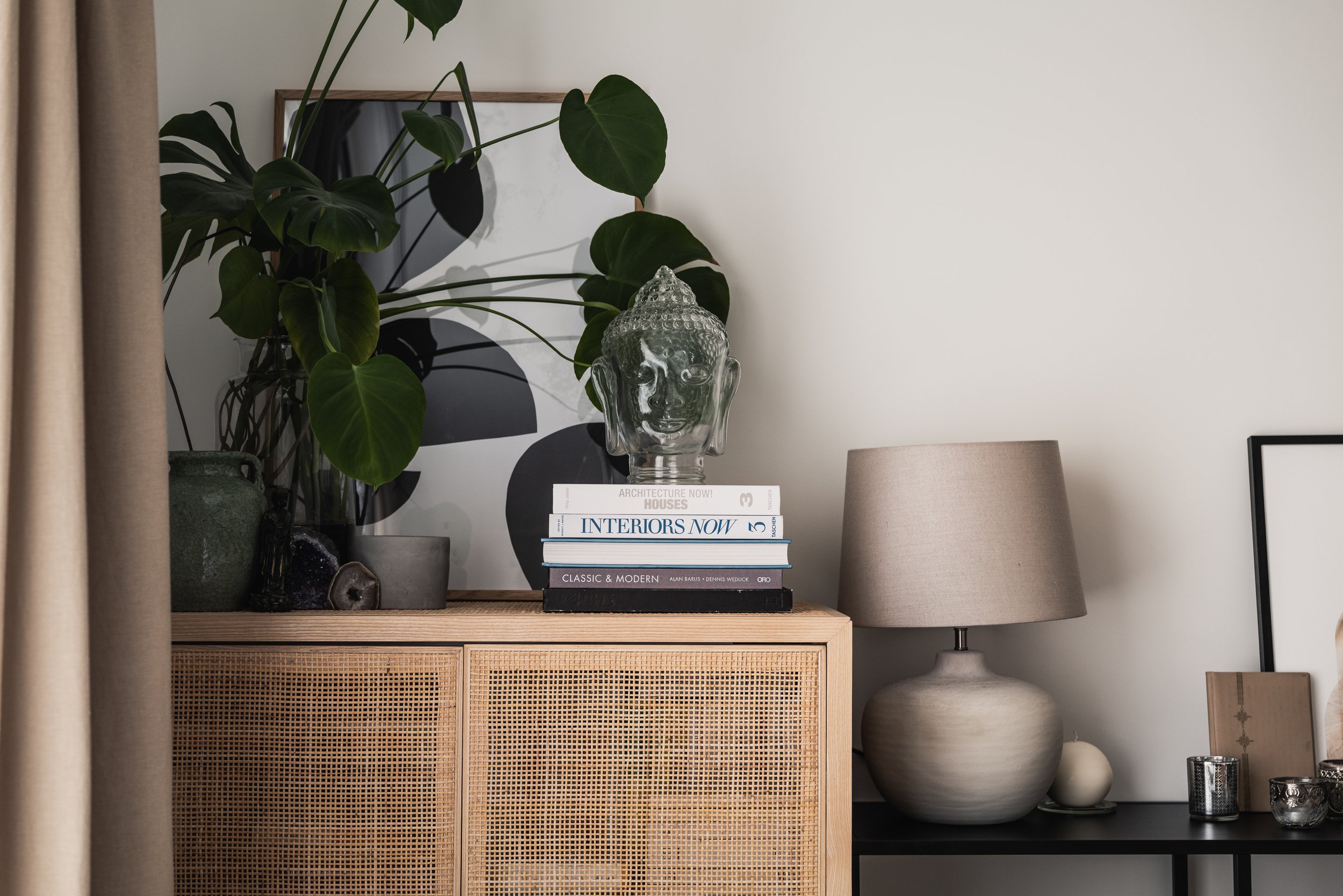 Interior decor scene with a rattan cabinet, potted plant, stacked books, a glass Buddha head sculpture, and a beige table lamp on a black side table.