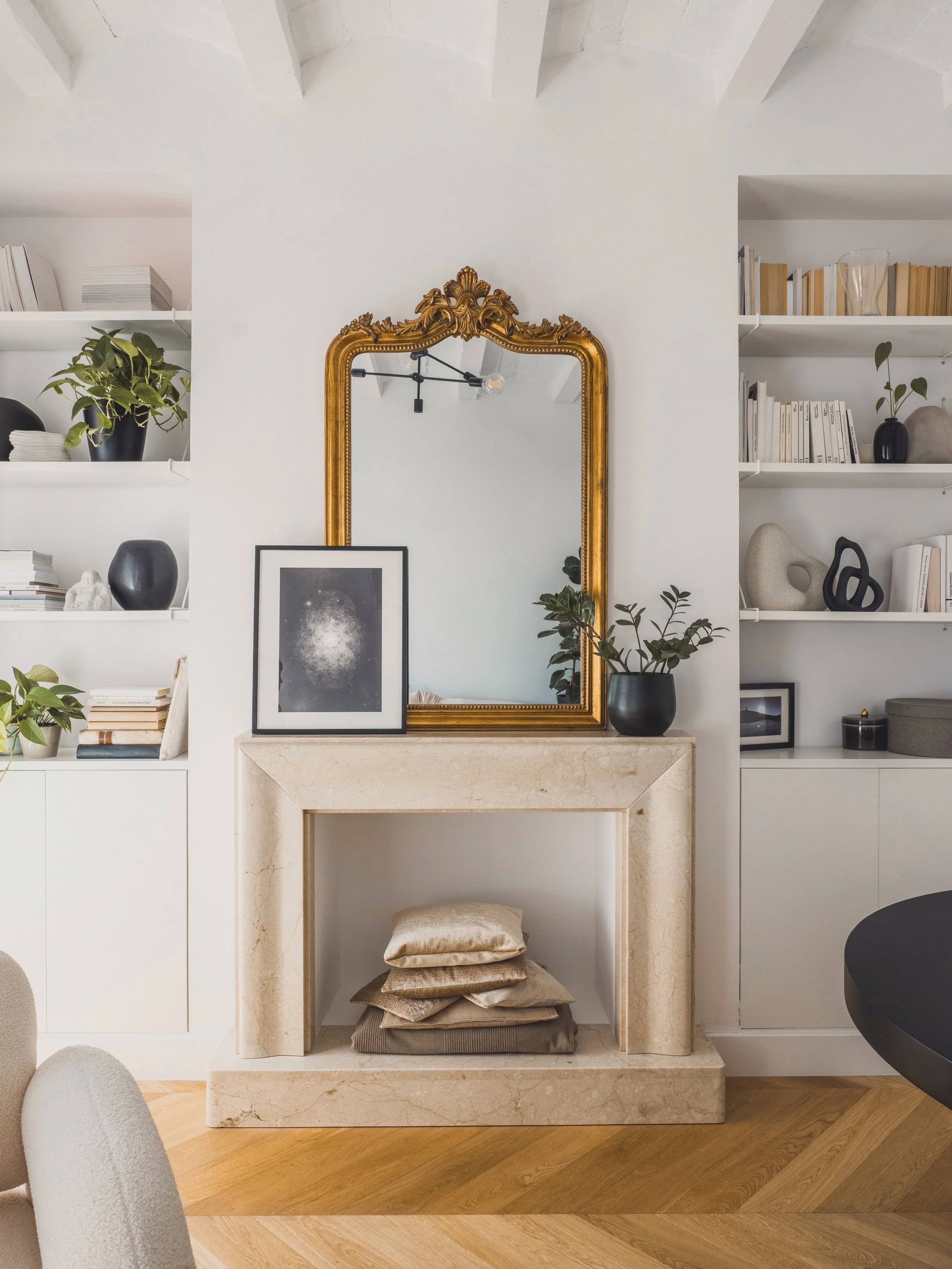 A living room with a white wall, an ornate gold-framed mirror, and built-in white shelves filled with books, decorative objects, and plants. There is a beige marble fireplace with stacked cushions and a tufted ottoman underneath.