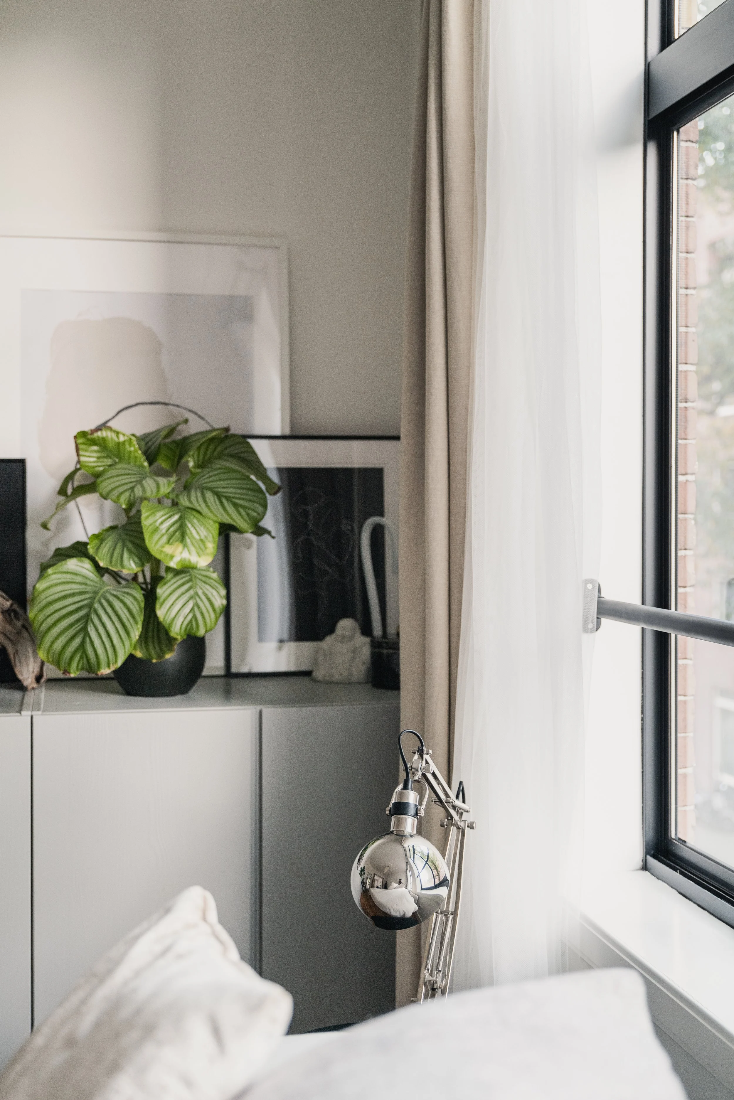 A window with sheer white curtains in a modern living room, decorated with framed artwork and potted plants on a white cabinet, with a silver adjustable desk lamp in the foreground.