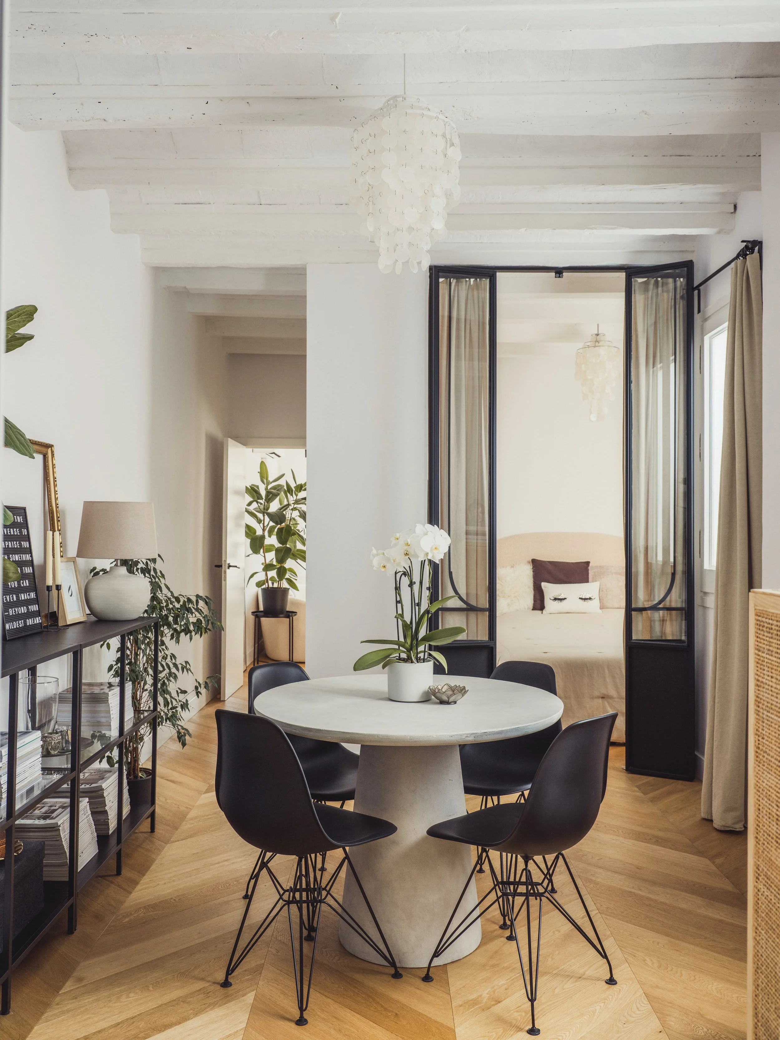 A modern dining room with black chairs around a round white table with a potted orchid. The room has a wooden floor, white walls, and a ceiling with exposed white beams. A black metal framed door leads to a cozy seating area with beige curtains and a