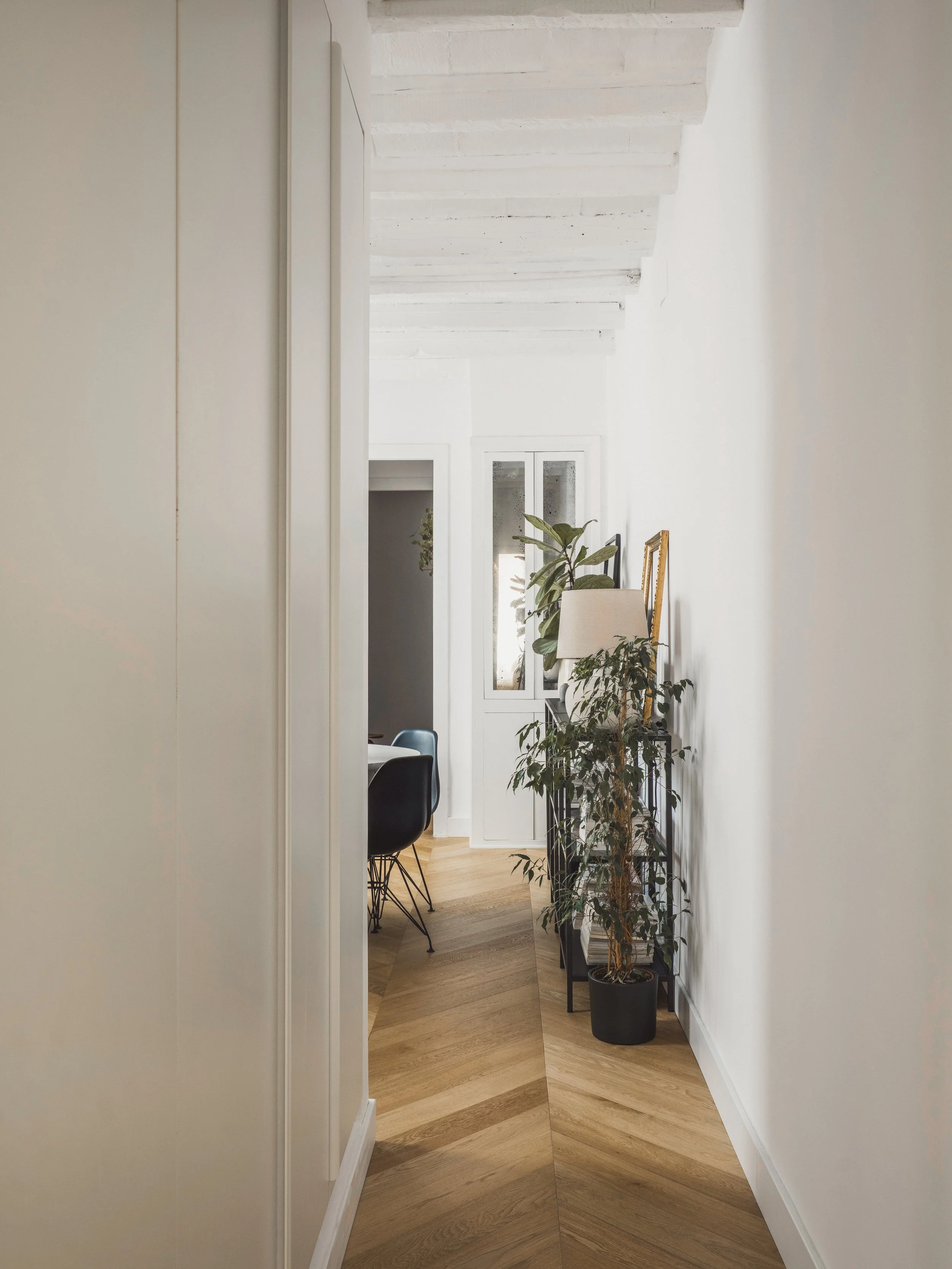 A narrow hallway leading to a room with a white cabinet, green plants, a beige lamp, and a black chair with a wooden table, with white walls and wooden flooring.