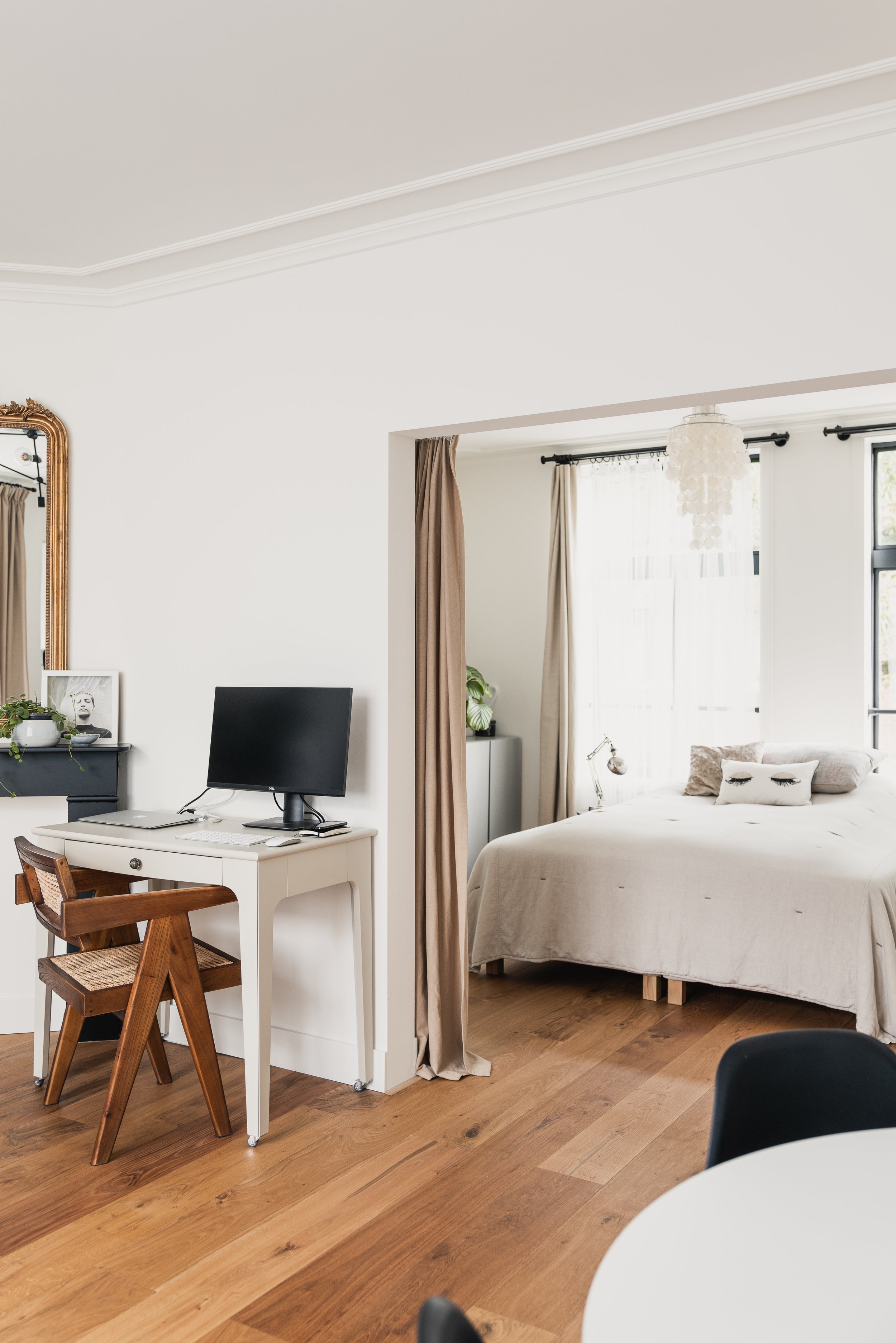 Modern bedroom with a white bed, beige pillows, and a window with curtains, separated by a curtain divider from a workspace with a small desk, computer, and wooden chair, in a bright and airy room with hardwood floors.
