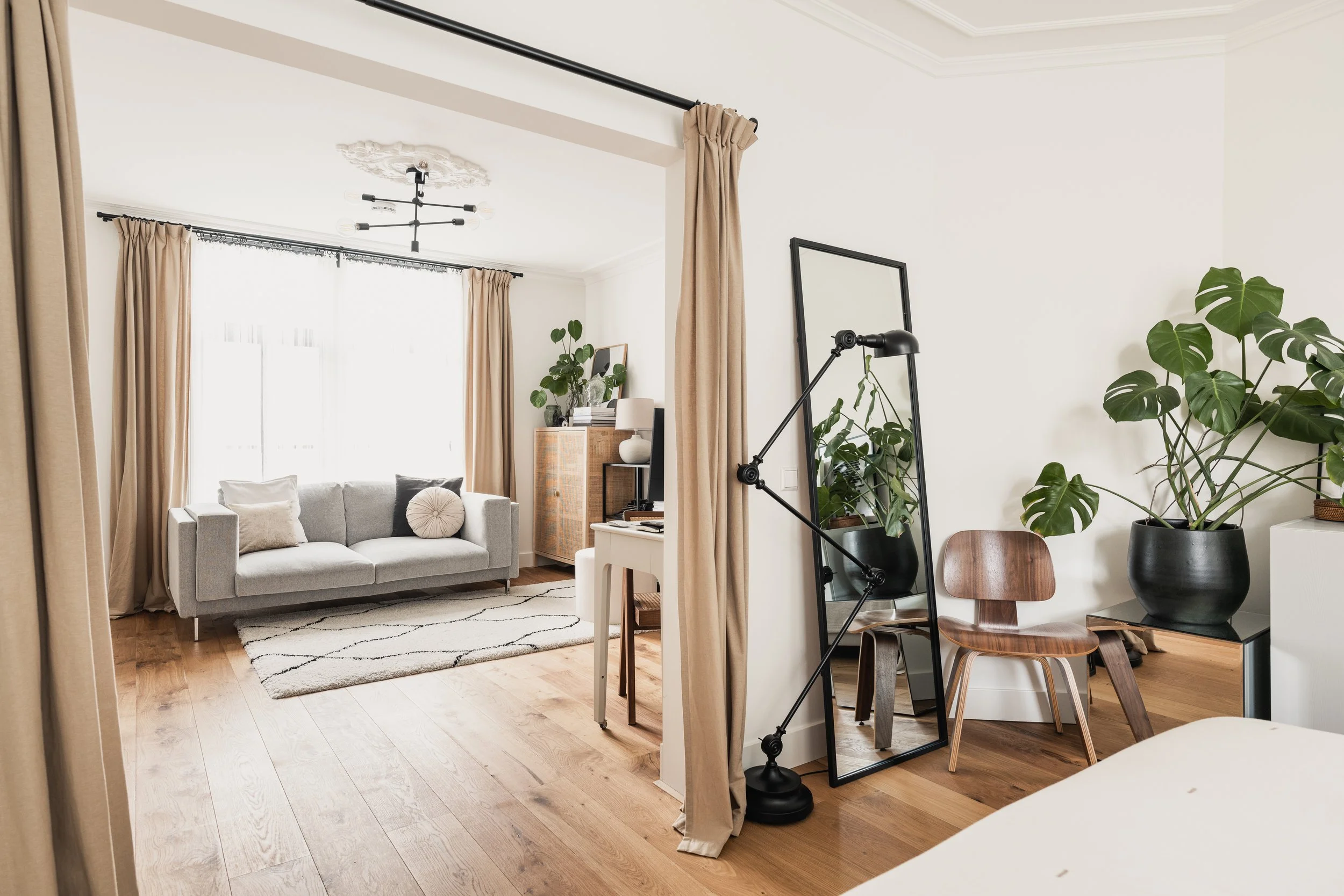 Living room with a grey sofa, a white desk with a chair, houseplants, a mirror, and beige curtains, with hardwood floors and natural light from a large window.
