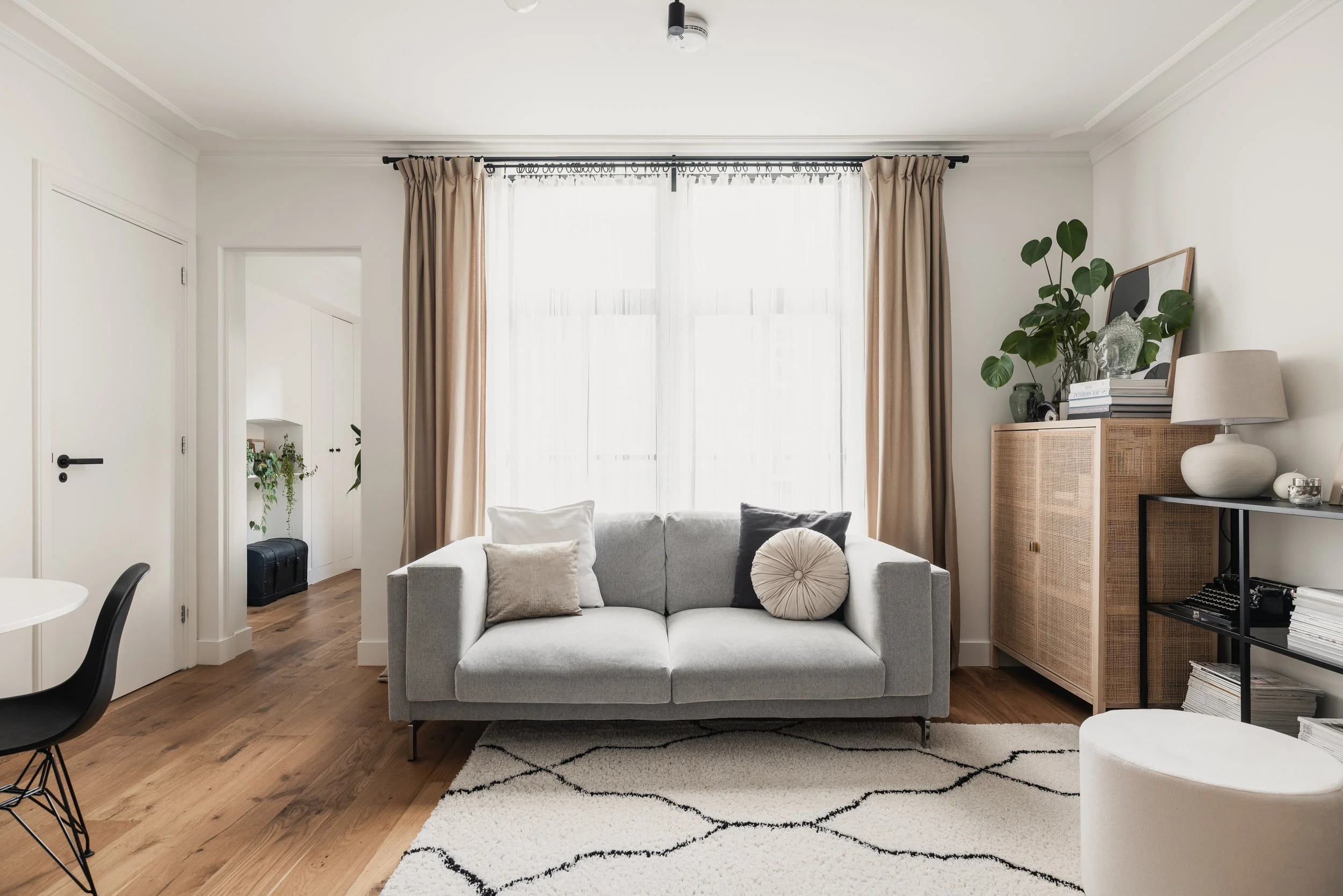 A bright living room with a gray sofa, decorative pillows, a patterned rug, and a window with beige curtains. There is a wooden cabinet with plants and artwork on top, and a black shelf with books and decor items. A white ottoman is in the foreground