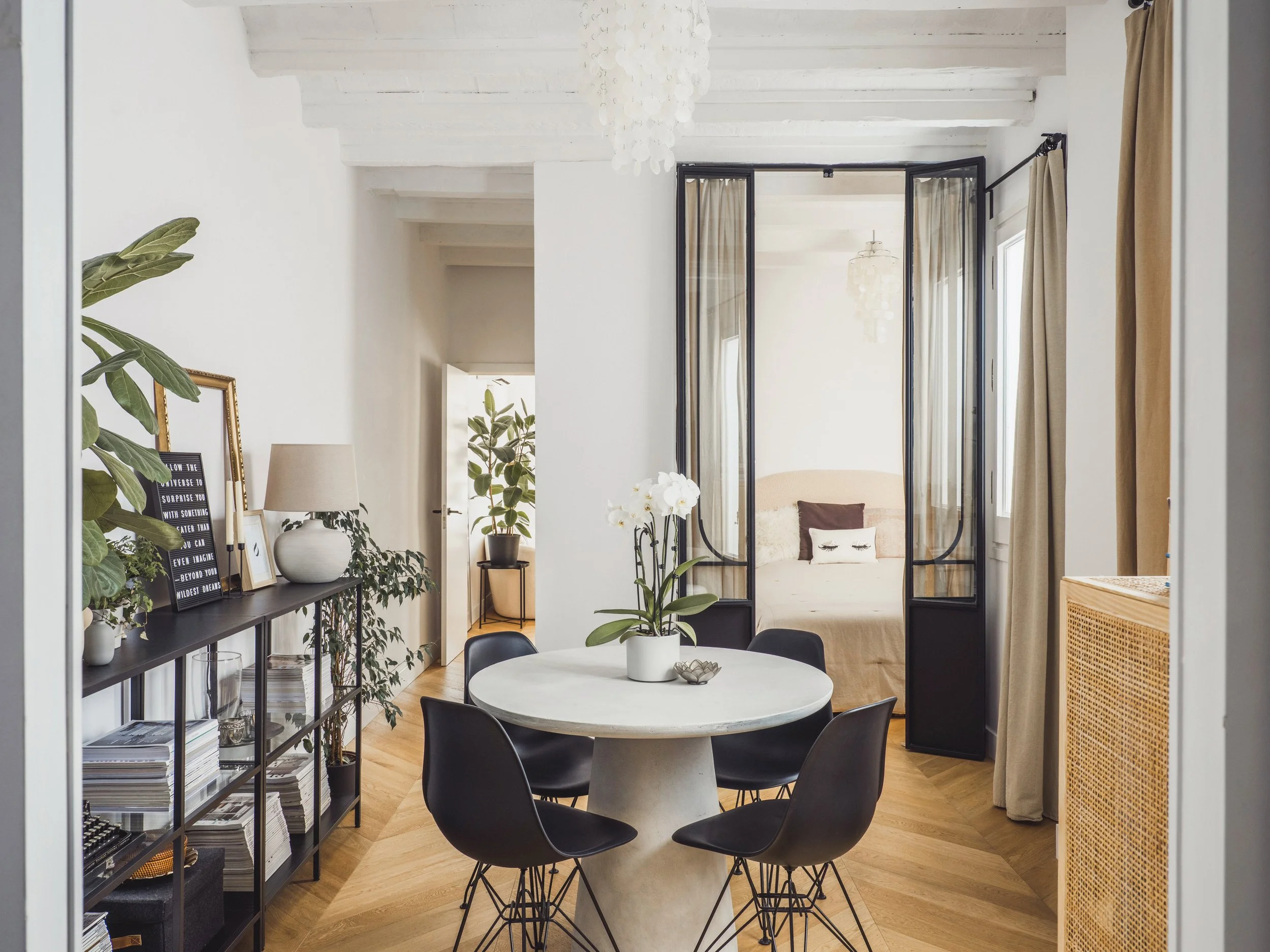 A dining area with a round white table, black chairs, and various decorative plants and picture frames. There is a black shelving unit with books and decorations along the left wall. The room has large windows with beige curtains, and a bed with pill
