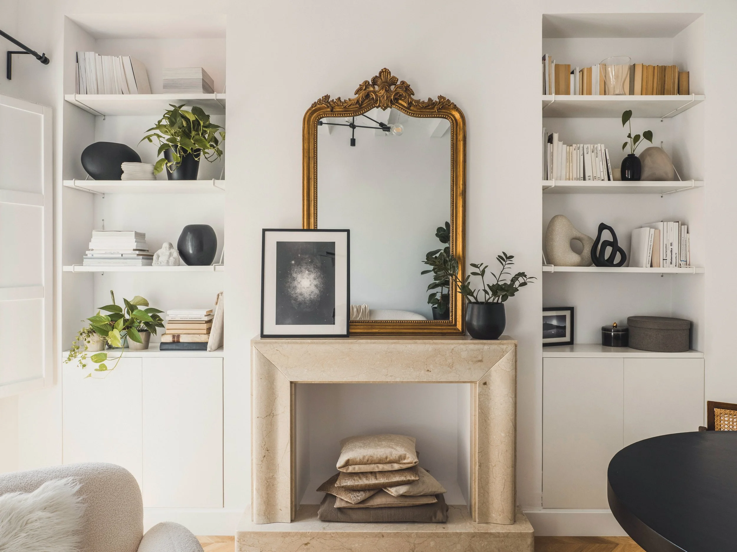 A built-in white bookcase with open shelves displaying books, decorative objects, and potted plants. A large gold-framed mirror is centered on the wall above a marble fireplace, with a collage of framed art and stacked cushions underneath.