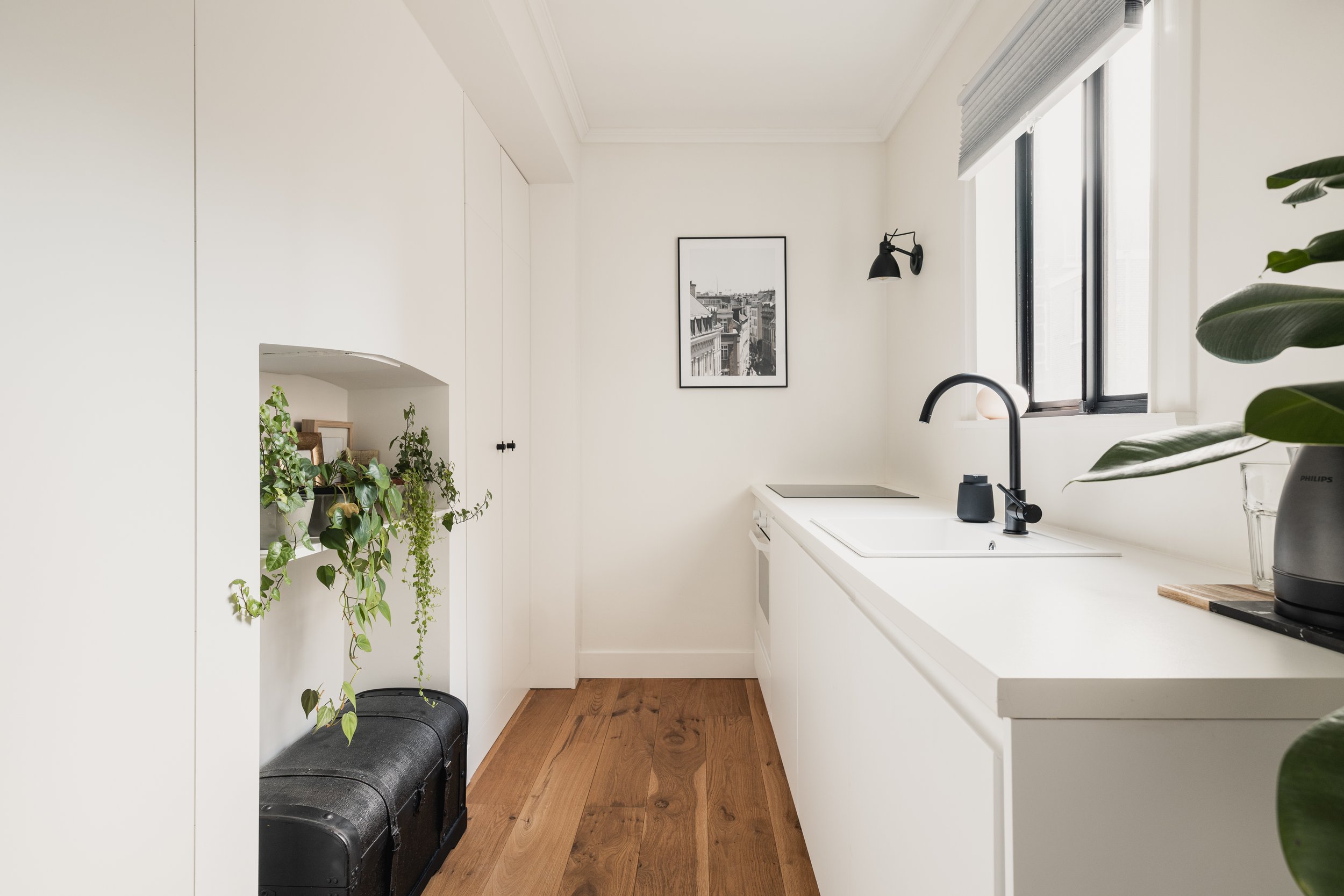 Minimalist white kitchen with black fixtures, wooden floor, and a window with gray blinds. Decor includes potted plants, a framed black and white photo, and a black kettle.