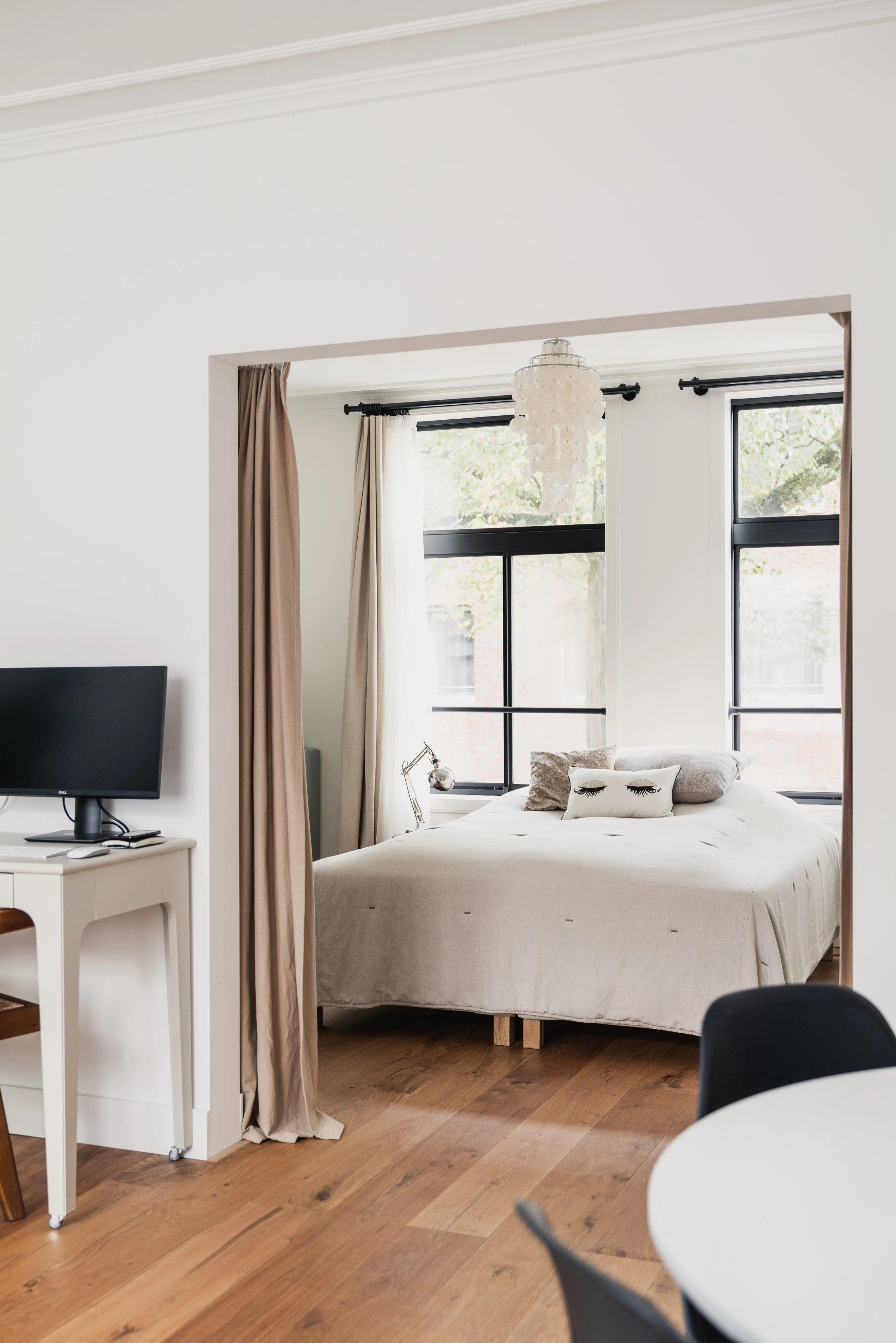 Modern bedroom with large windows, white walls, beige bed, decorative pillows, bedside lamp, and a small TV on a white desk