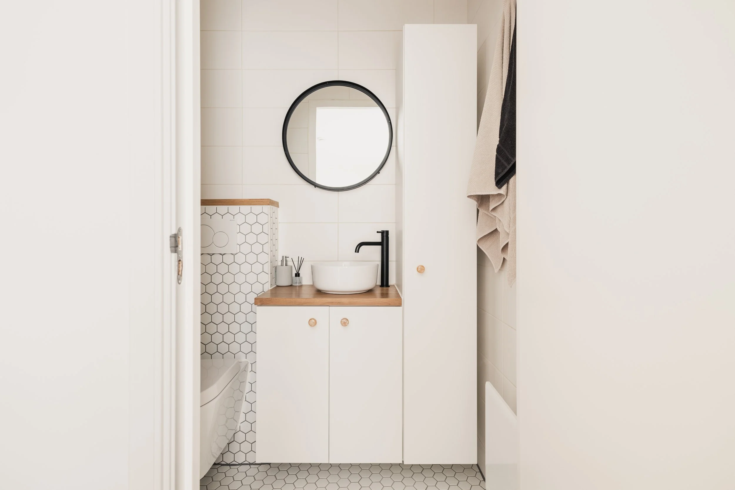 Minimalist bathroom with a round mirror, black faucet, white vessel sink, wood countertop, and white cabinets. Towel hanging on the wall, patterned hexagon tile on the wall and floor.