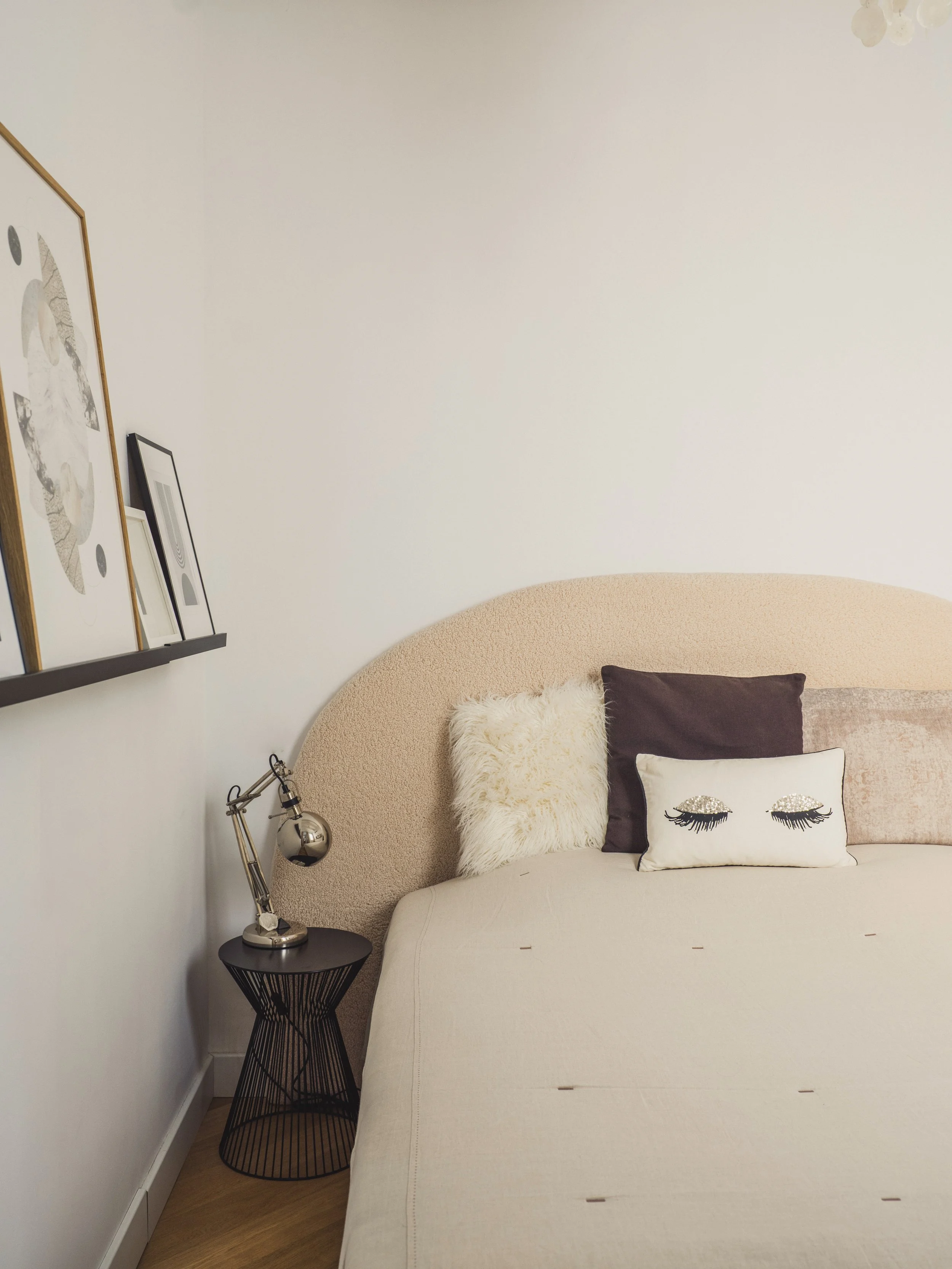 A bedroom corner with a beige bed, decorative pillows including one with closed eyes makeup design, a black wire side table, a modern desk lamp, and framed artwork on a white wall.