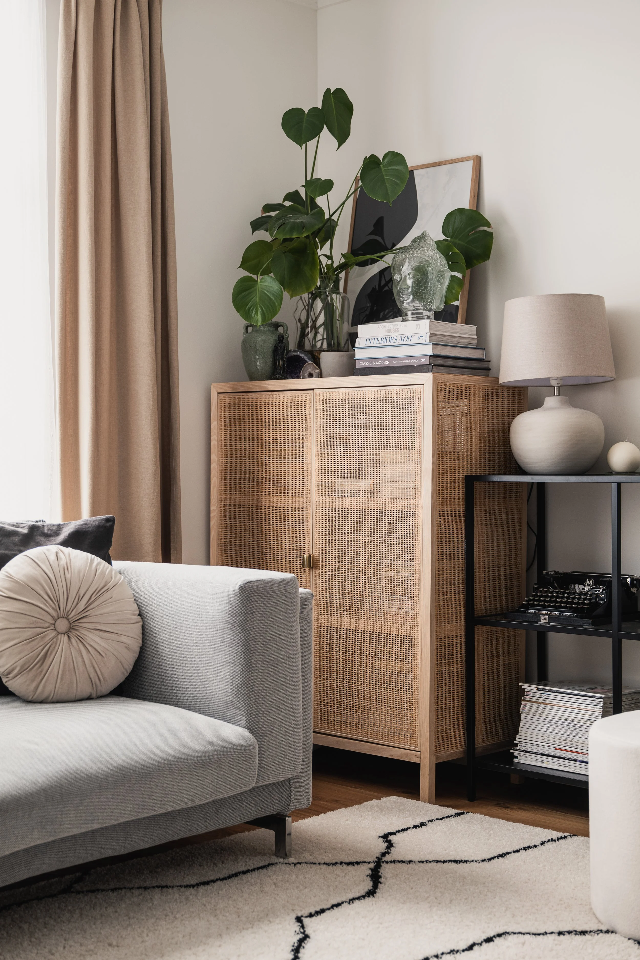 Living room with light gray sofa, round beige pillow, beige curtains, wooden cabinet topped with large green plant, books, and decorative items, black metal shelf with vintage radio and magazines, beige lamp with round ceramic base, and white wall.